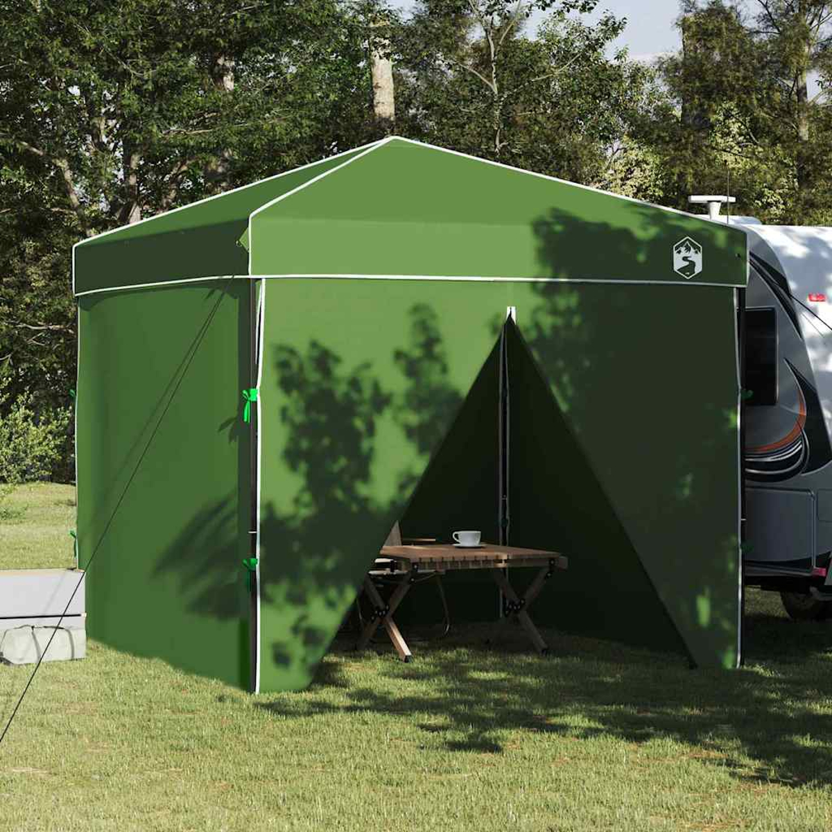 Un rifugio da campeggio verde con un tavolo e una tazza all'interno. Un camper sullo sfondo. Alberi ed erba circondano il rifugio.
