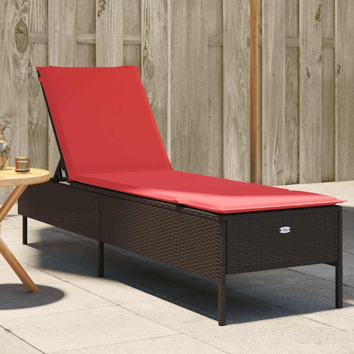 Eine rot gepolsterte Sonnenliege steht auf einer Terrasse. Ein kleiner Tisch und ein Holzzaun befinden sich im Hintergrund.