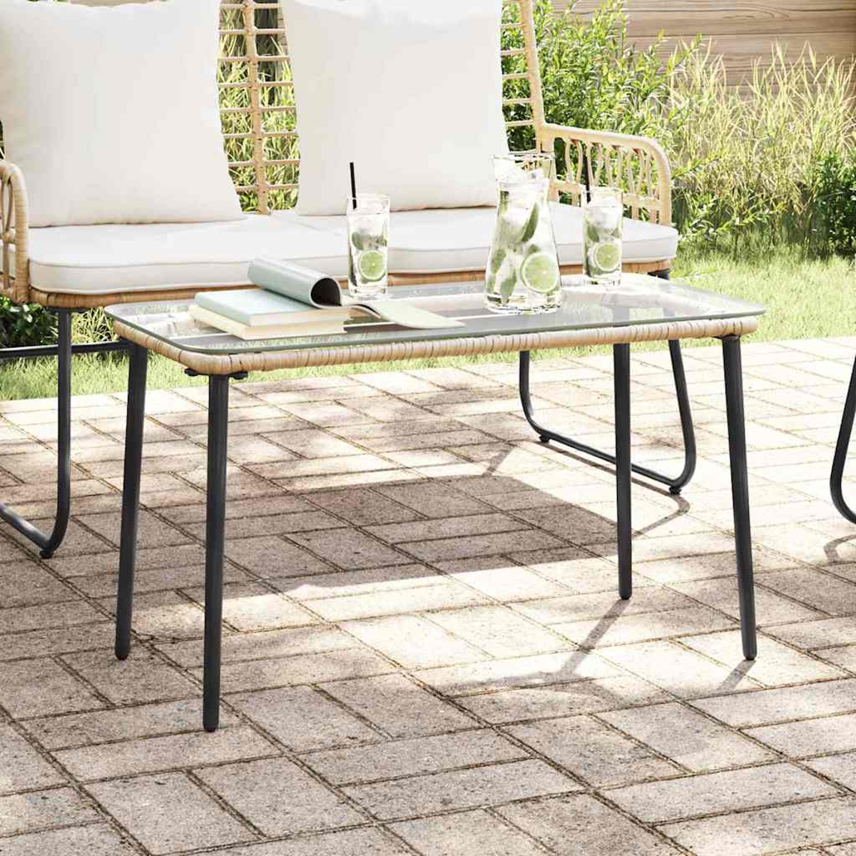 Escena al aire libre: mesa de centro con tapa de cristal con bebidas, libros y una silla de mimbre con cojines blancos en un patio de ladrillos.