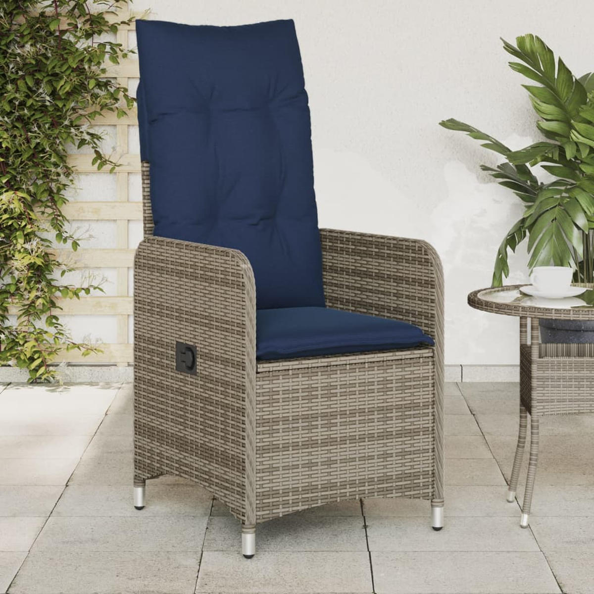 Silla de mimbre con cojín azul. A la derecha, una mesita con una taza. Plantas verdes y una pared blanca al fondo.