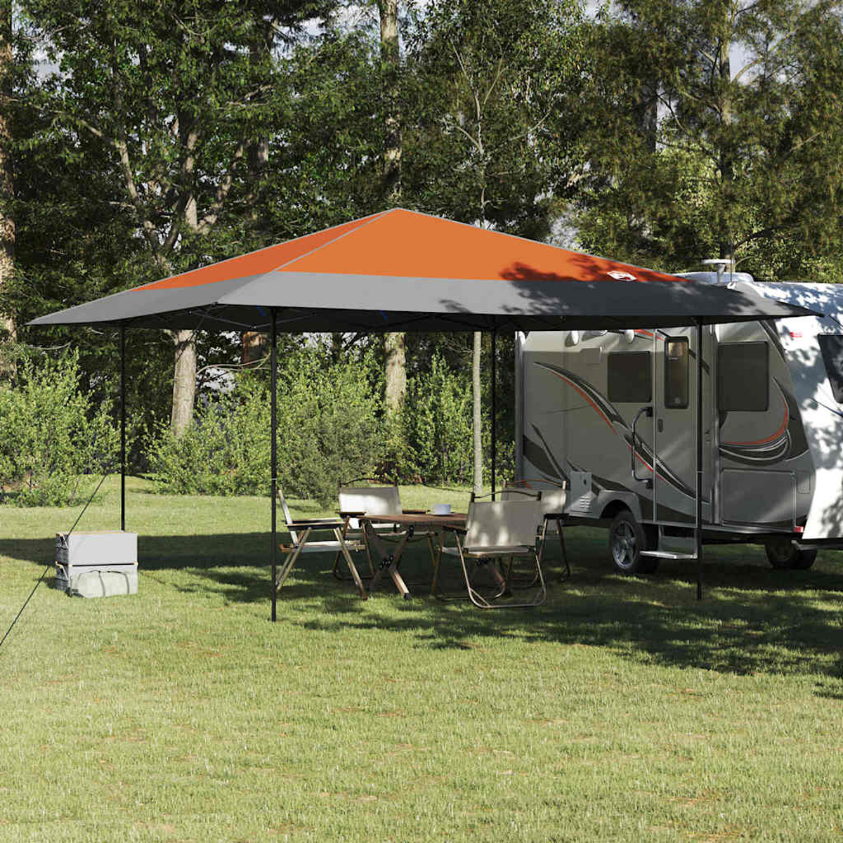 Una tenda arancione e grigia sopra un tavolo e sedie, un camper e alberi sull'erba.