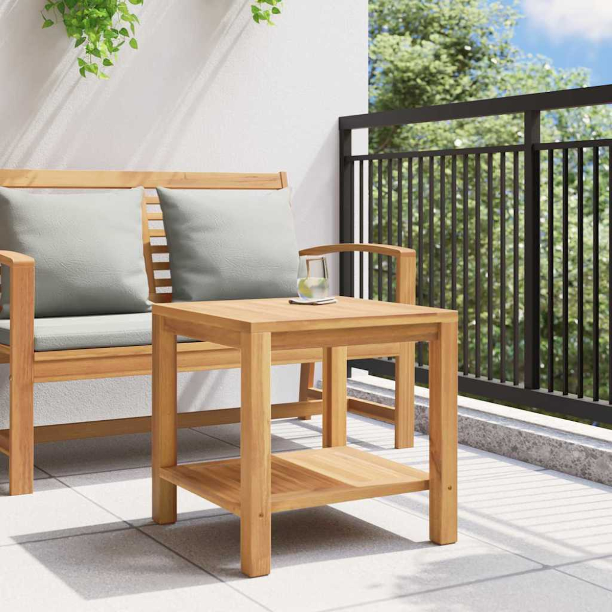 Ein Holztisch mit einem Glas Wasser auf einer Terrasse. Dahinter steht eine Bank mit Kissen. Ein schwarzes Geländer und Bäume sind im Hintergrund.