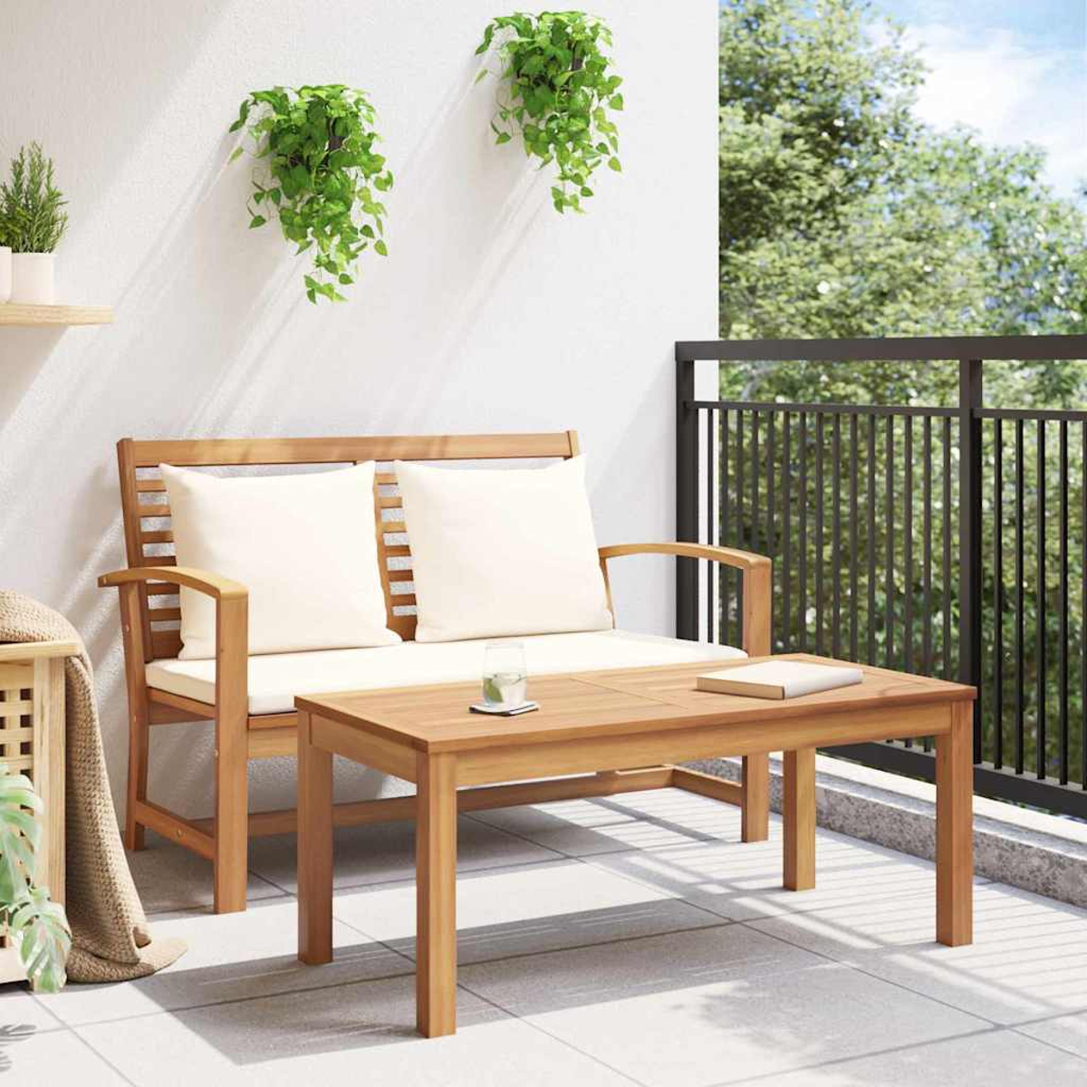 Holzbank und Tisch auf dem Balkon. Weiße Kissen auf der Bank. Grüne Pflanzen hängen an der Wand. Glas und Buch liegen auf dem Tisch.