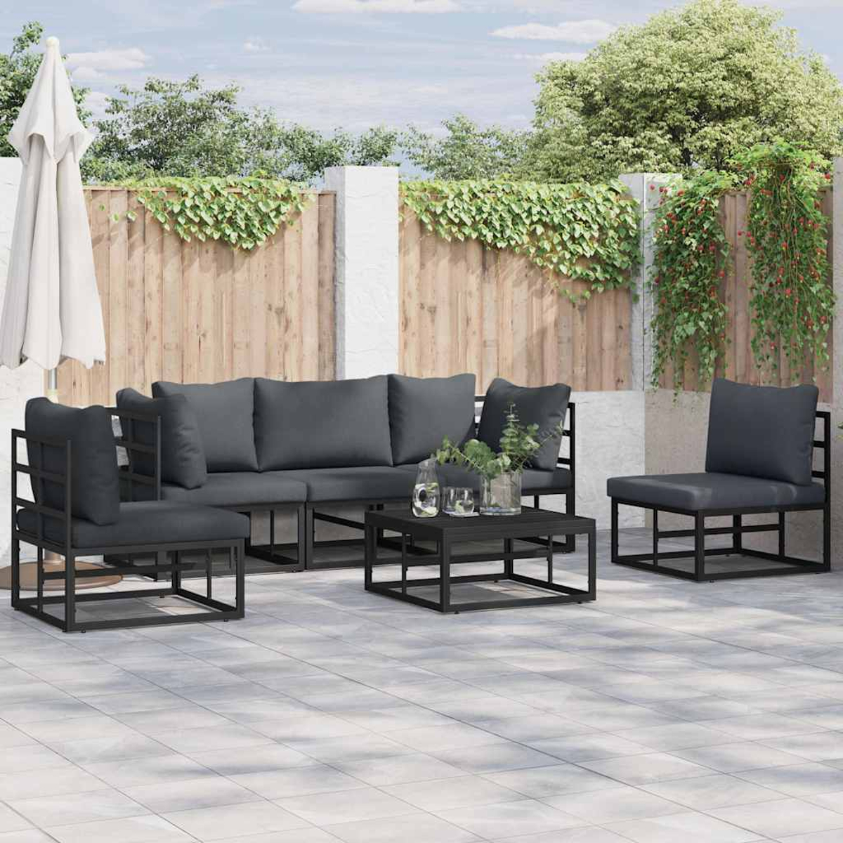 Set di mobili da patio: cuscini grigi, telai neri, tavolino e ombrellone bianco. Staccionata in legno con piante.
