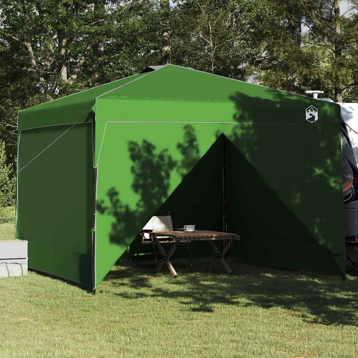 Riparo da campeggio verde con parte anteriore aperta, tavolo, tazza, sedia. Alberi sullo sfondo, camper parcheggiato. Erba in primo piano.