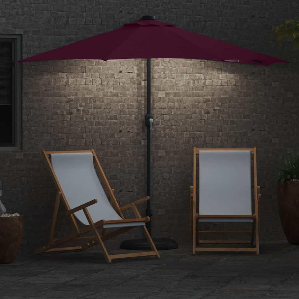 Un ombrello bordeaux aperto si trova sopra due sedie a sdraio in legno su un patio in mattoni. Un muro di mattoni fa da sfondo.
