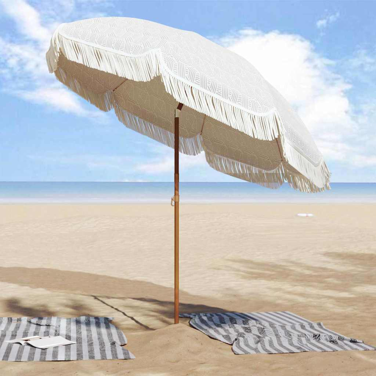 Un ombrellone da spiaggia bianco con frange su una spiaggia sabbiosa. Due teli da spiaggia a righe e l'oceano sono visibili sullo sfondo.