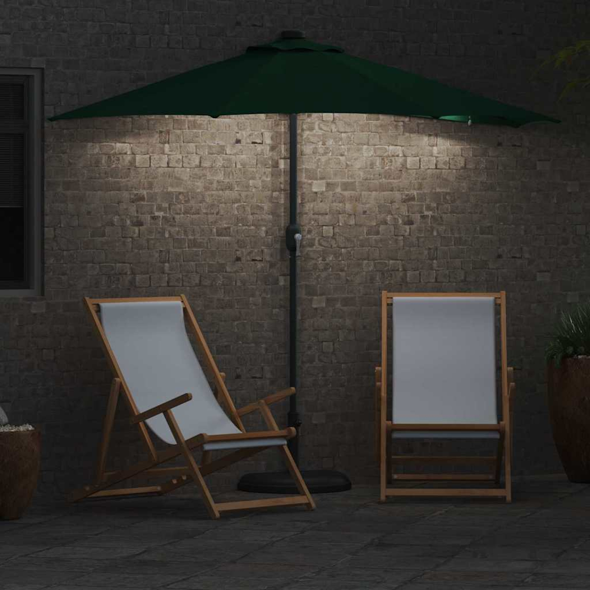Una scena di patio notturna con un ombrellone verde, due sedie bianche e un muro di mattoni illuminato da una lampada.