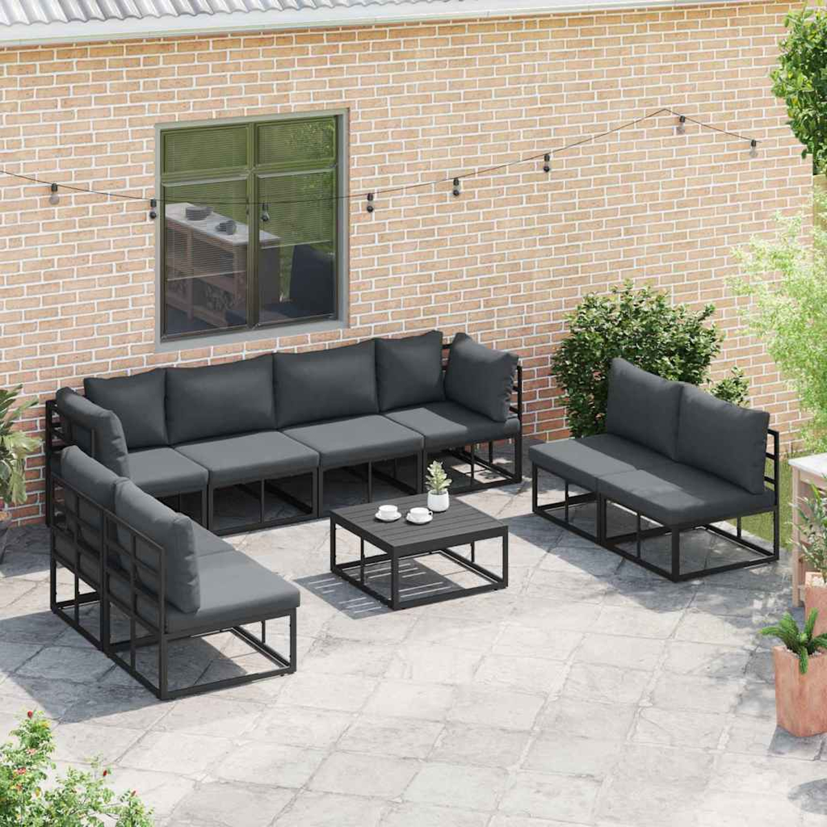 Outdoor-Sitzbereich mit grauen Kissen und schwarzen Rahmen. Ein kleiner Tisch mit Tassen steht in der Mitte. Ziegelmauer im Hintergrund.