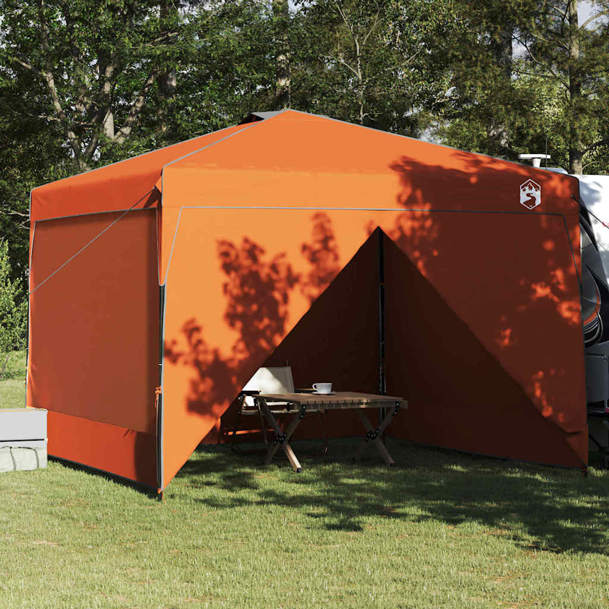 Ein orangefarbener Campingunterstand auf einer Rasenfläche. Ein Tisch, Stühle und eine Tasse sind drinnen. Bäume bilden den Hintergrund.