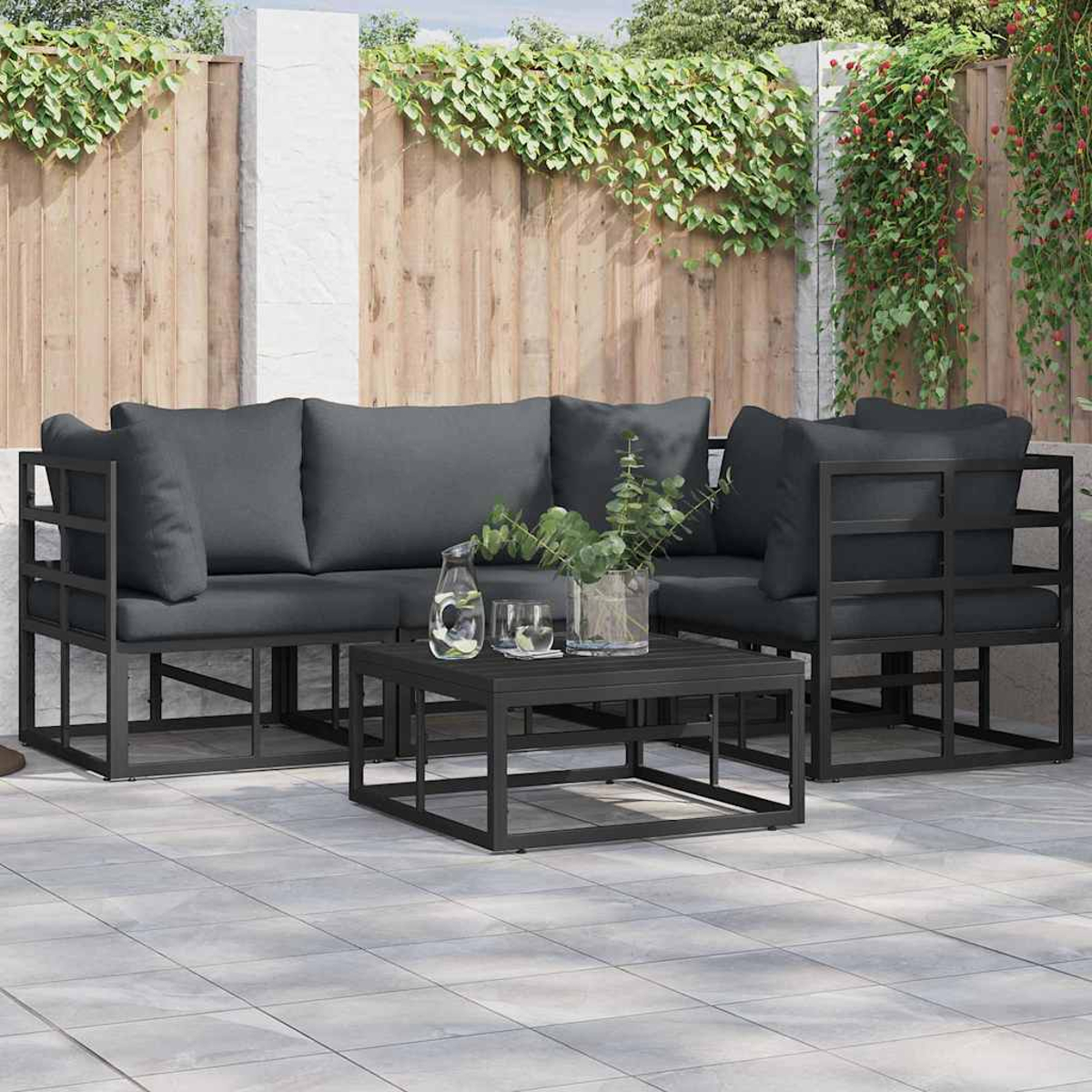 Outdoor-Ecksofa mit grauen Kissen und schwarzem Metallrahmen. Davor stehen ein schwarzer Tisch und Pflanzen. Holzzaun im Hintergrund.