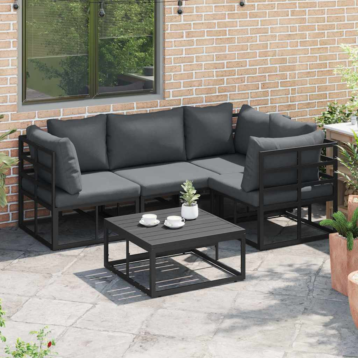 Gartenmöbel: graue Kissen, schwarze Rahmen, kleiner Tisch mit Tassen, Backsteinmauer im Hintergrund, Topfpflanzen.