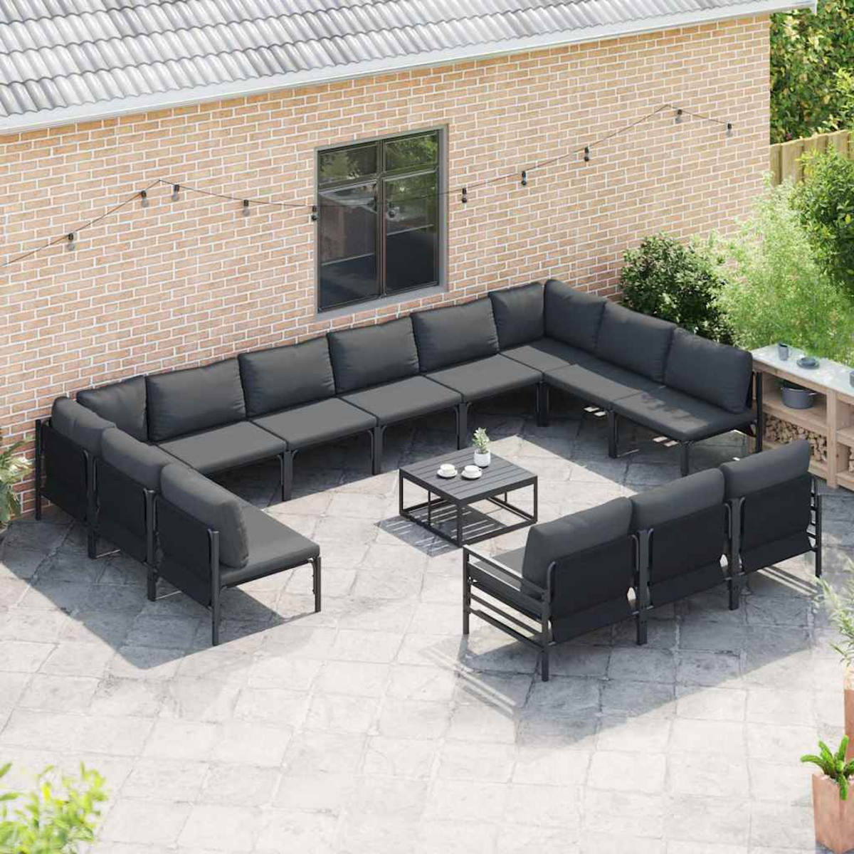 Terrassenmöbel-Set mit grauen Kissen. Um einen schwarzen quadratischen Tisch angeordnet. Hintergrund Backsteinmauer mit Fenster und grünen Pflanzen.