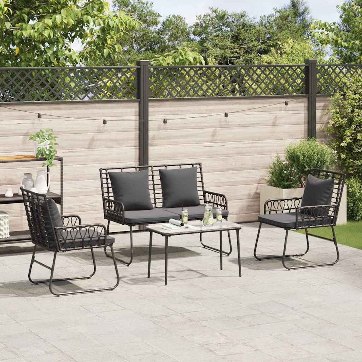 Juego de patio al aire libre con sillas de metal negro, un sofá y una mesa con cojines grises. Una valla de madera y vegetación al fondo.