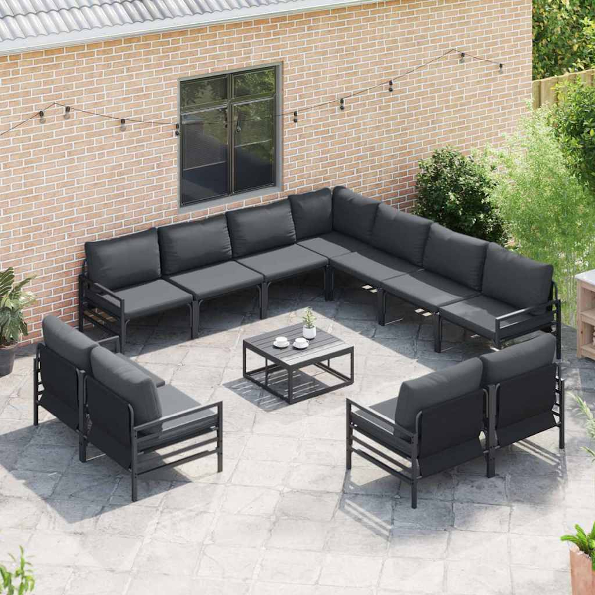 Outdoor-Sitzarrangement mit grauen Sofas und Sesseln um einen niedrigen Tisch, auf einer Terrasse mit Backsteinmauer im Hintergrund.