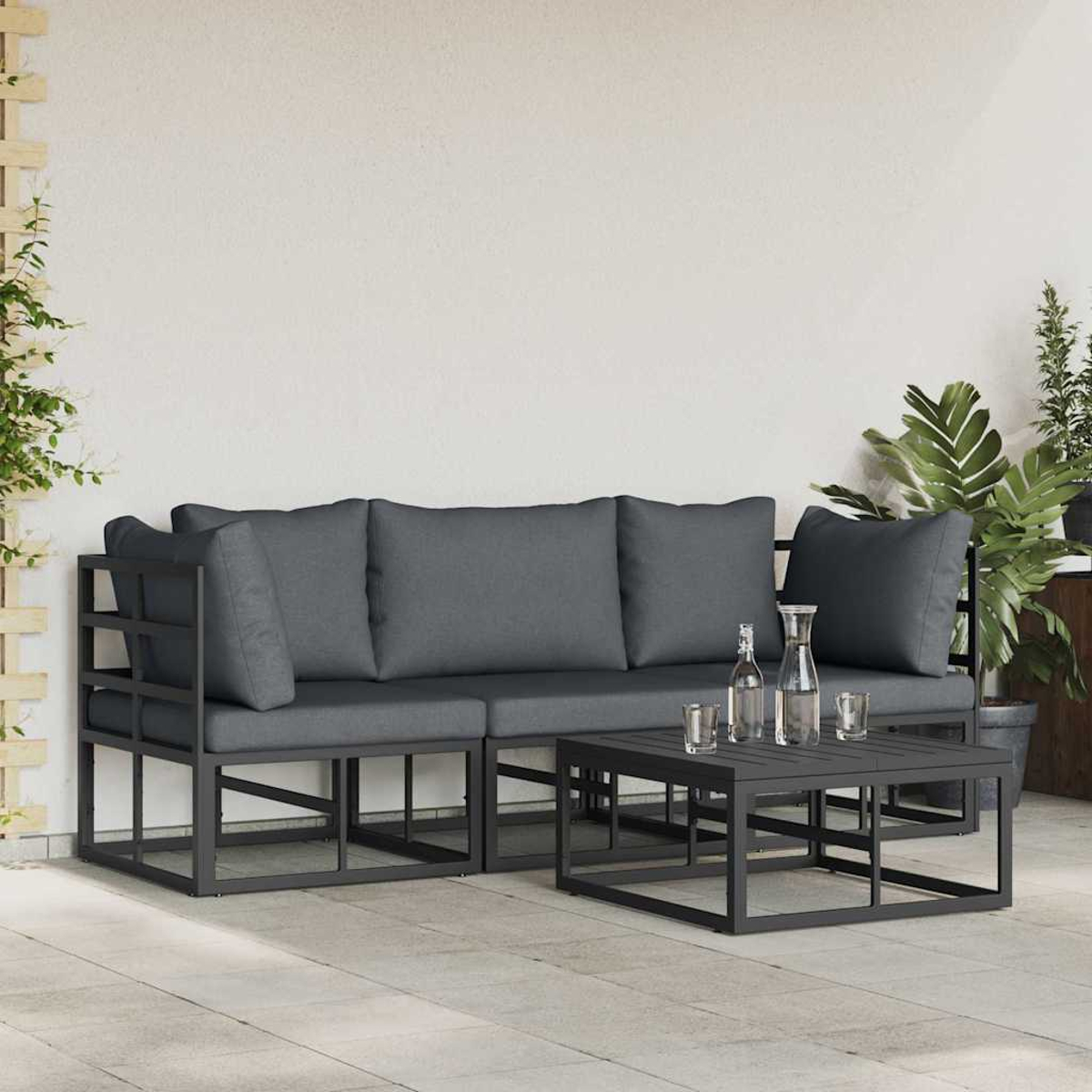 Set divani da esterno con cuscini grigi, tavolo quadrato con acqua e piante sullo sfondo. Muro bianco e pavimento piastrellato.