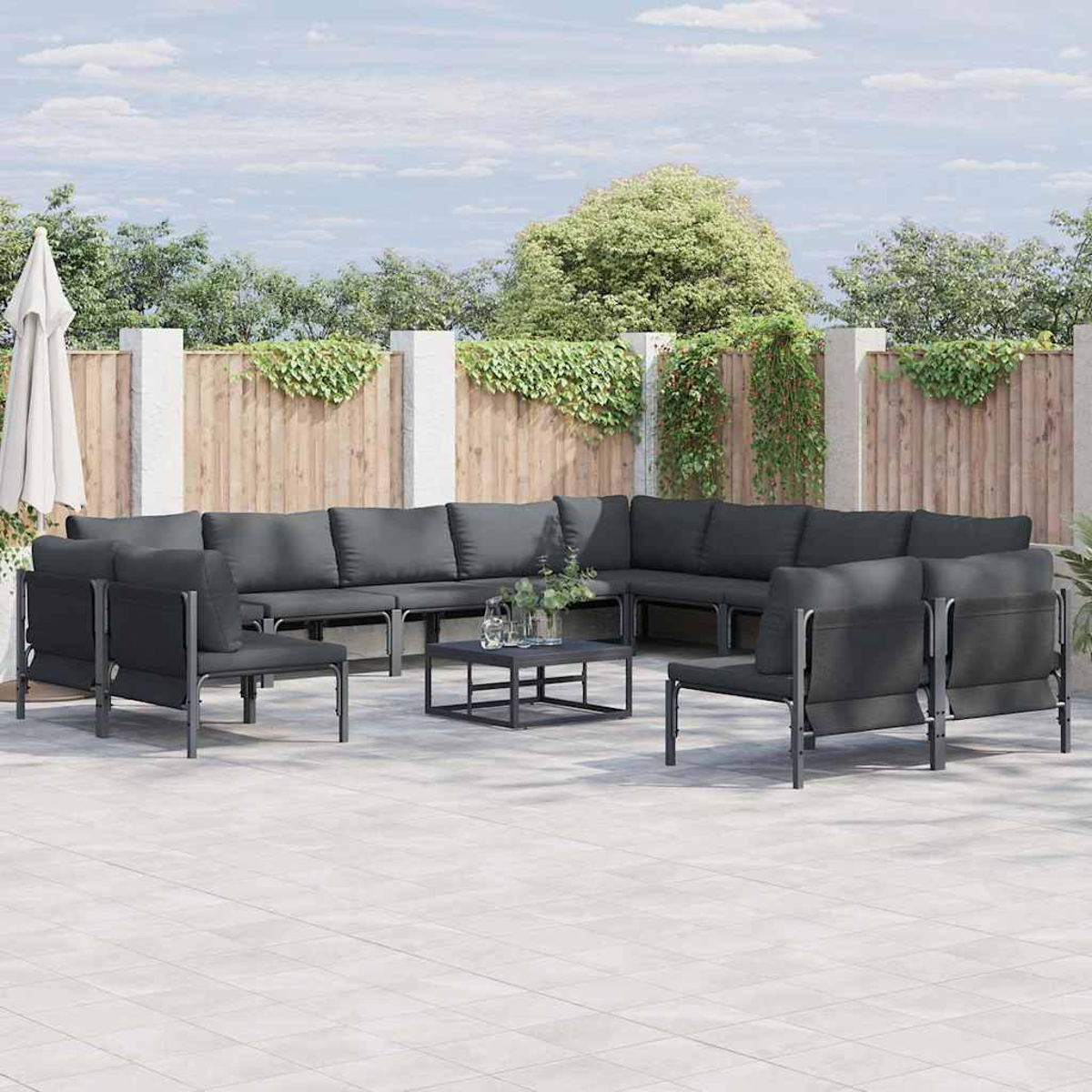 Outdoor-Sitzbereich mit grauen Kissen, einem schwarzen Tisch und einem weißen Sonnenschirm. Holzzaun, grüne Pflanzen und blauer Himmel im Hintergrund.