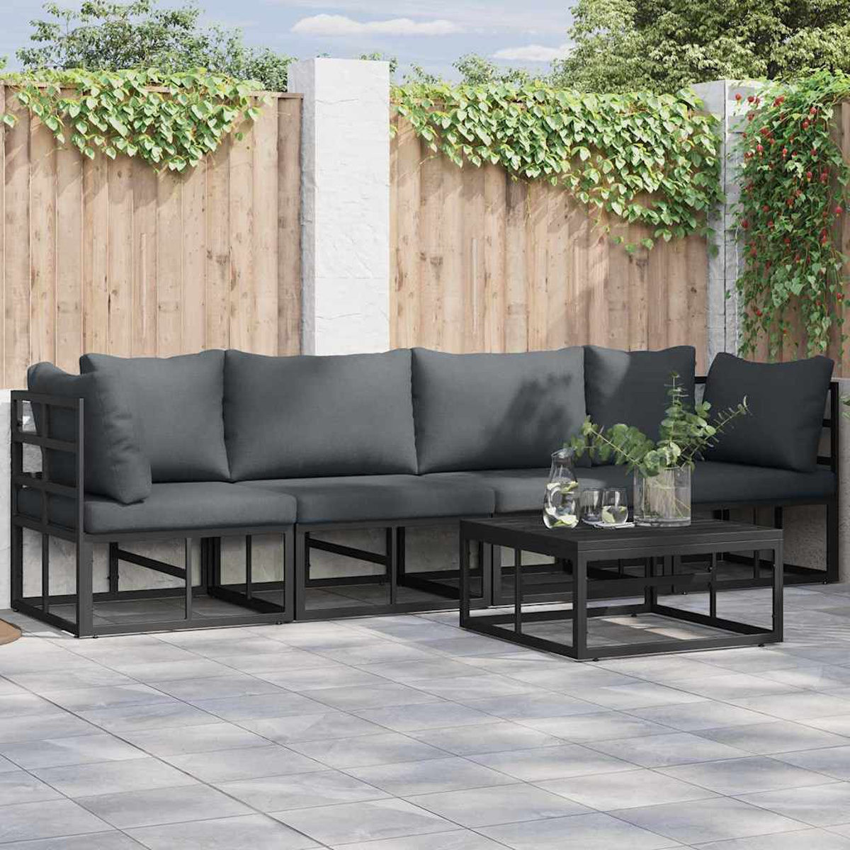 Terrassenset mit grauen Kissen, schwarzem Rahmen und kleinem Tisch. Grüne Pflanzen und Holzzaun im Hintergrund.