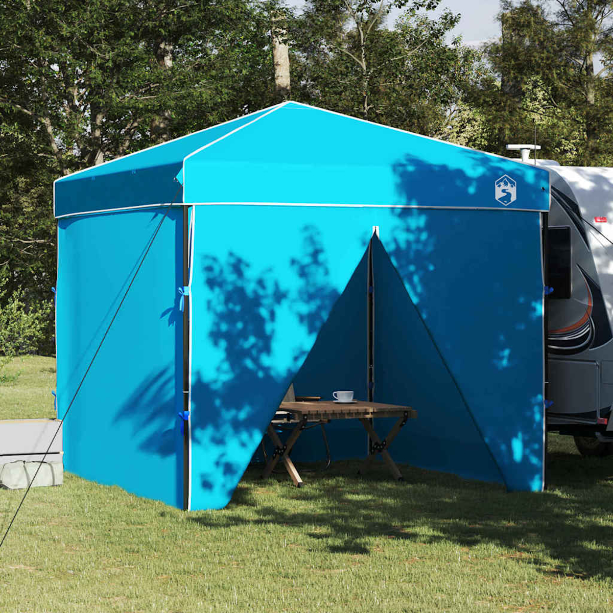 Una tenda blu con finiture bianche è allestita su un'area erbosa. Un tavolo e una roulotte sono visibili, con alberi sullo sfondo.