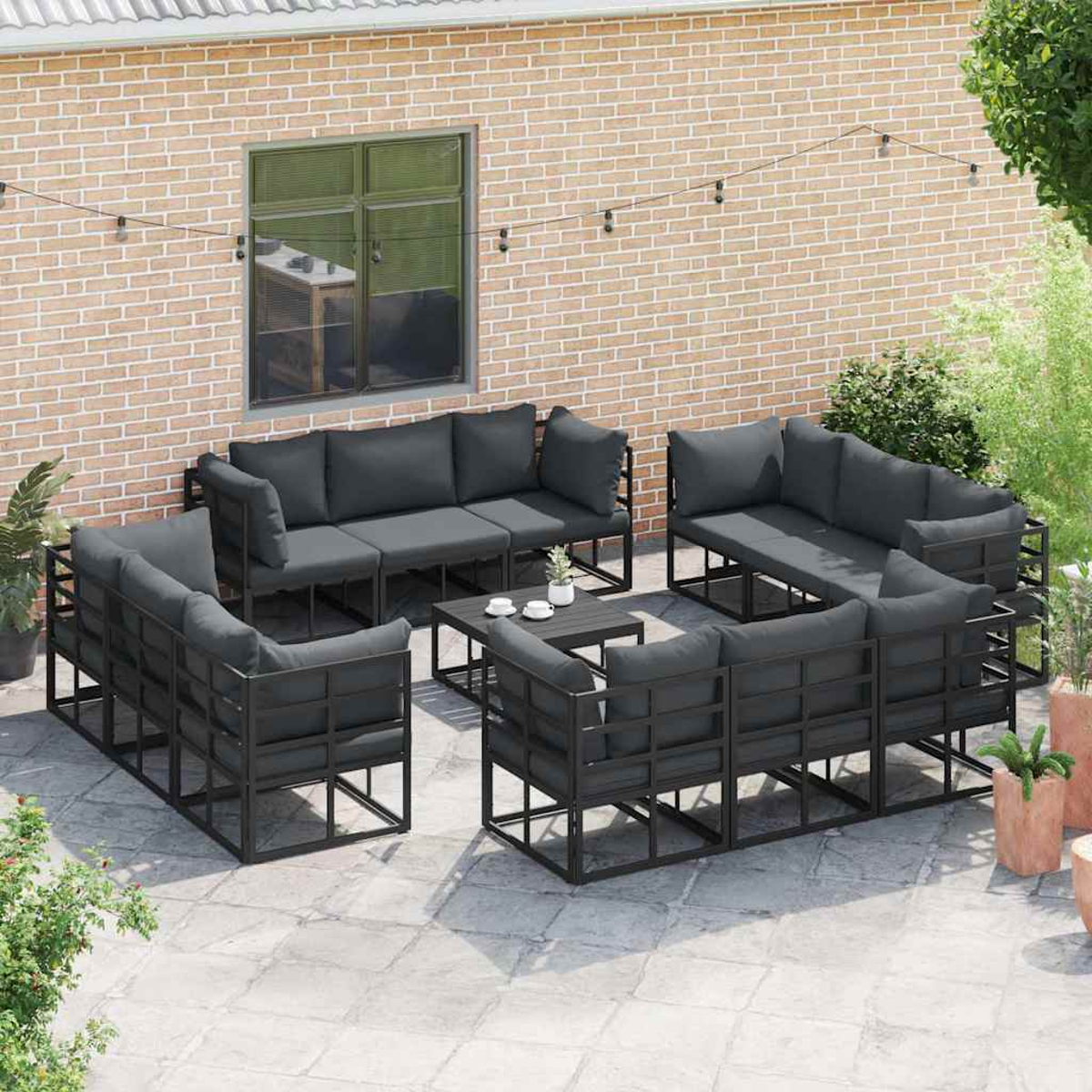 Set di mobili da patio esterno con cuscini grigi e struttura in metallo nero. Sfondo muro di mattoni, finestra e luci.