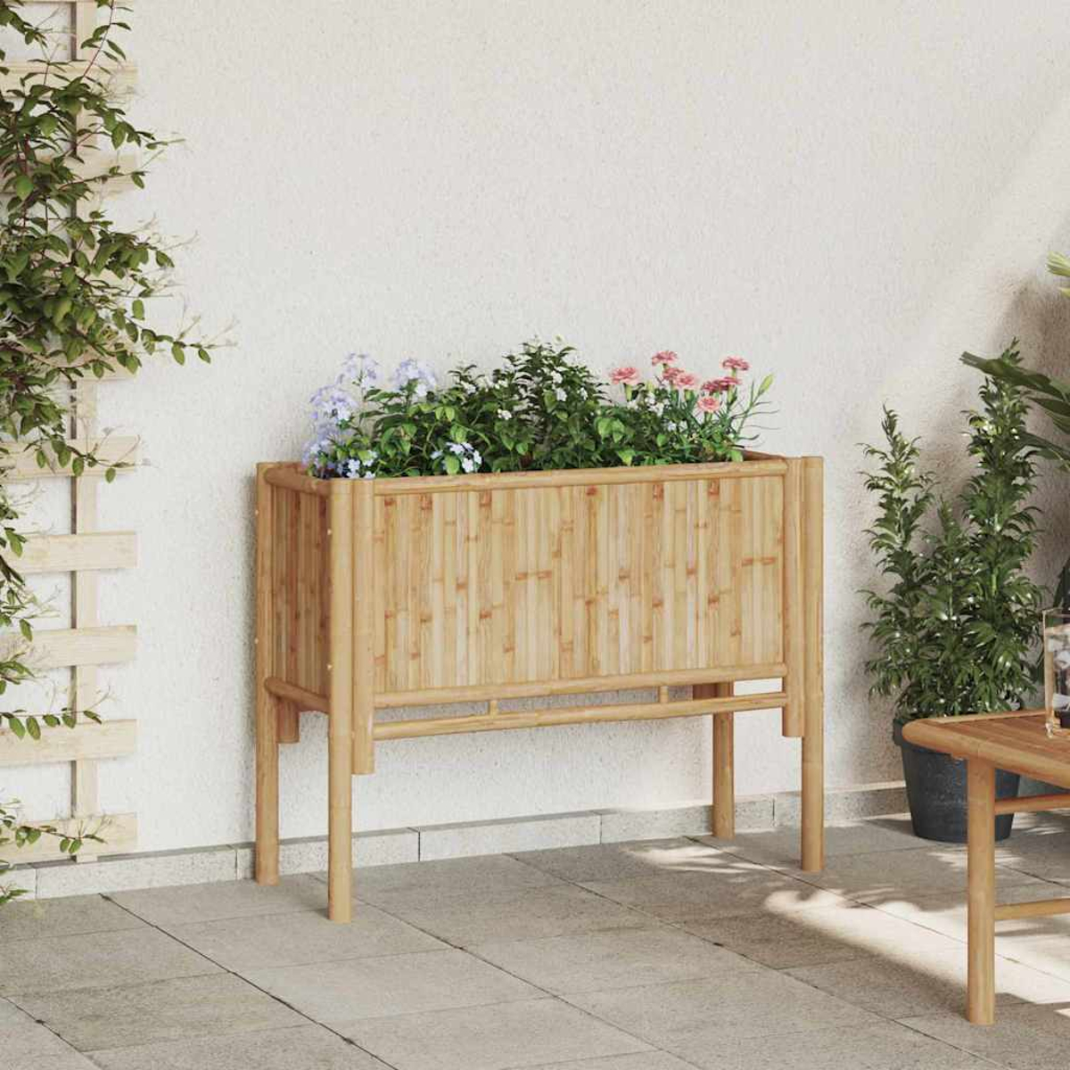 Una fioriera in bambù con fiori si trova su un patio. Una pianta rampicante cresce sul muro. Altre piante e un tavolo sono nelle vicinanze.