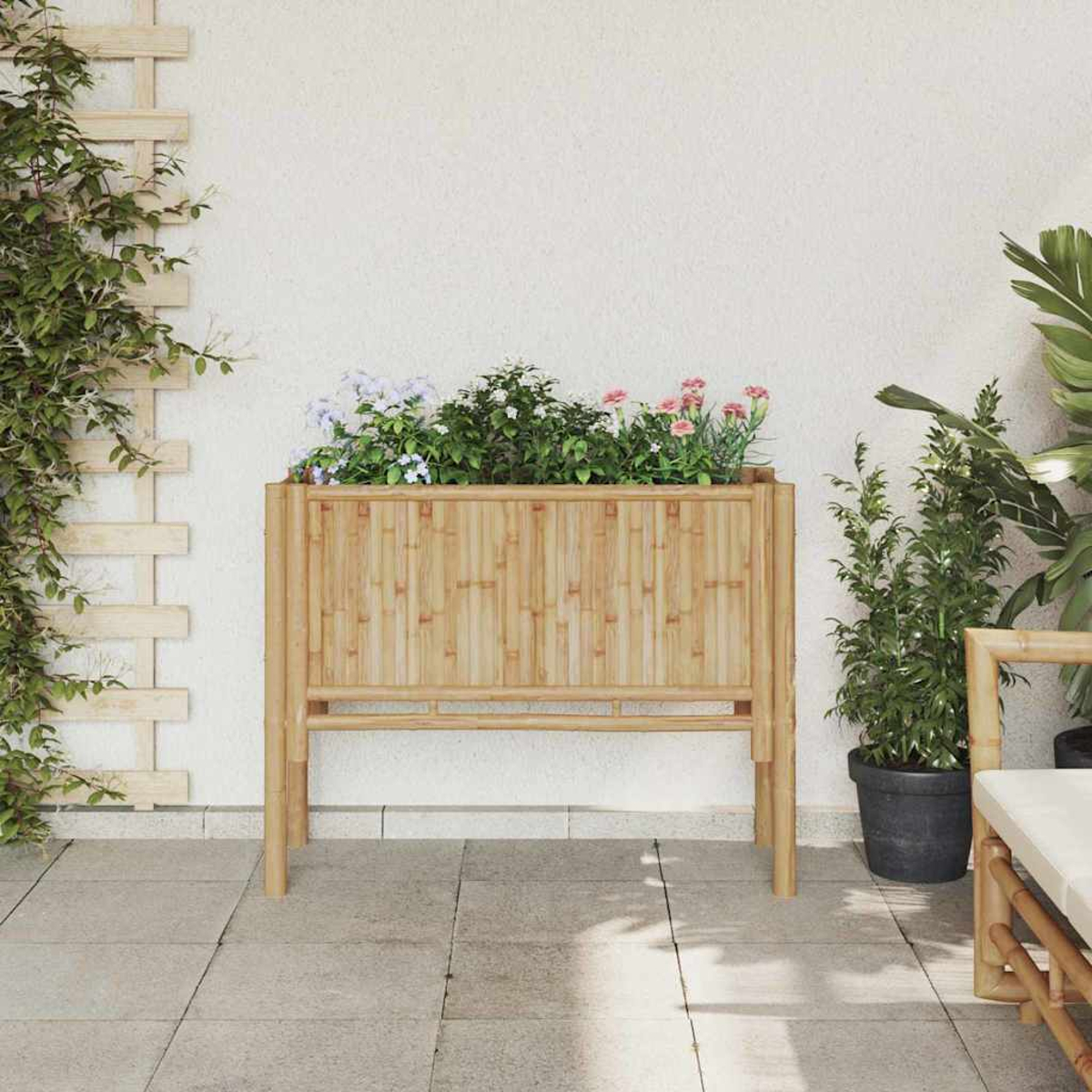 Una fioriera in bambù con fiori è su un patio. Un traliccio di legno con viti a sinistra. Piante e una sedia a destra.