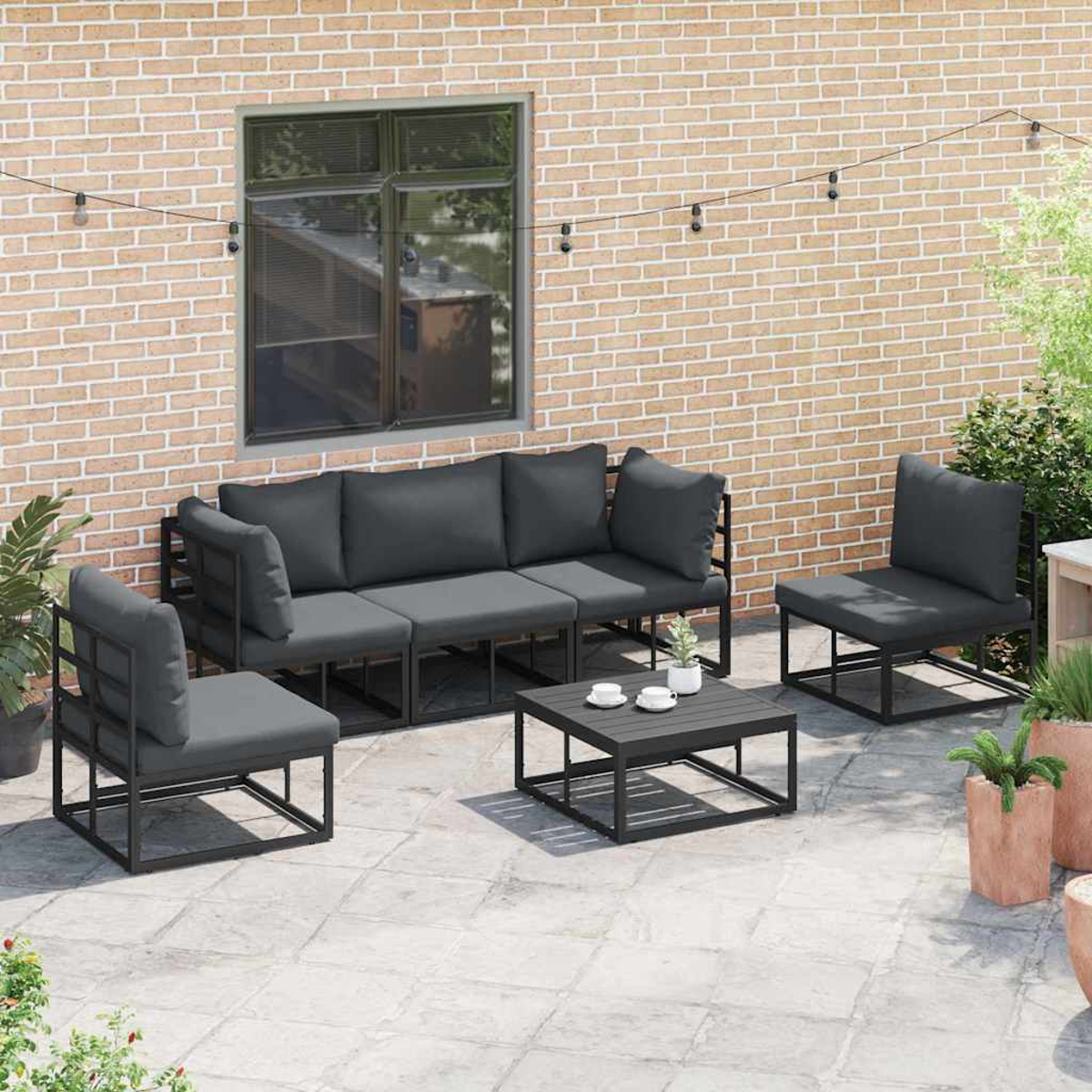 Set patio esterno con divano grigio, due sedie e tavolino. Contro un muro di mattoni con finestra e piante.