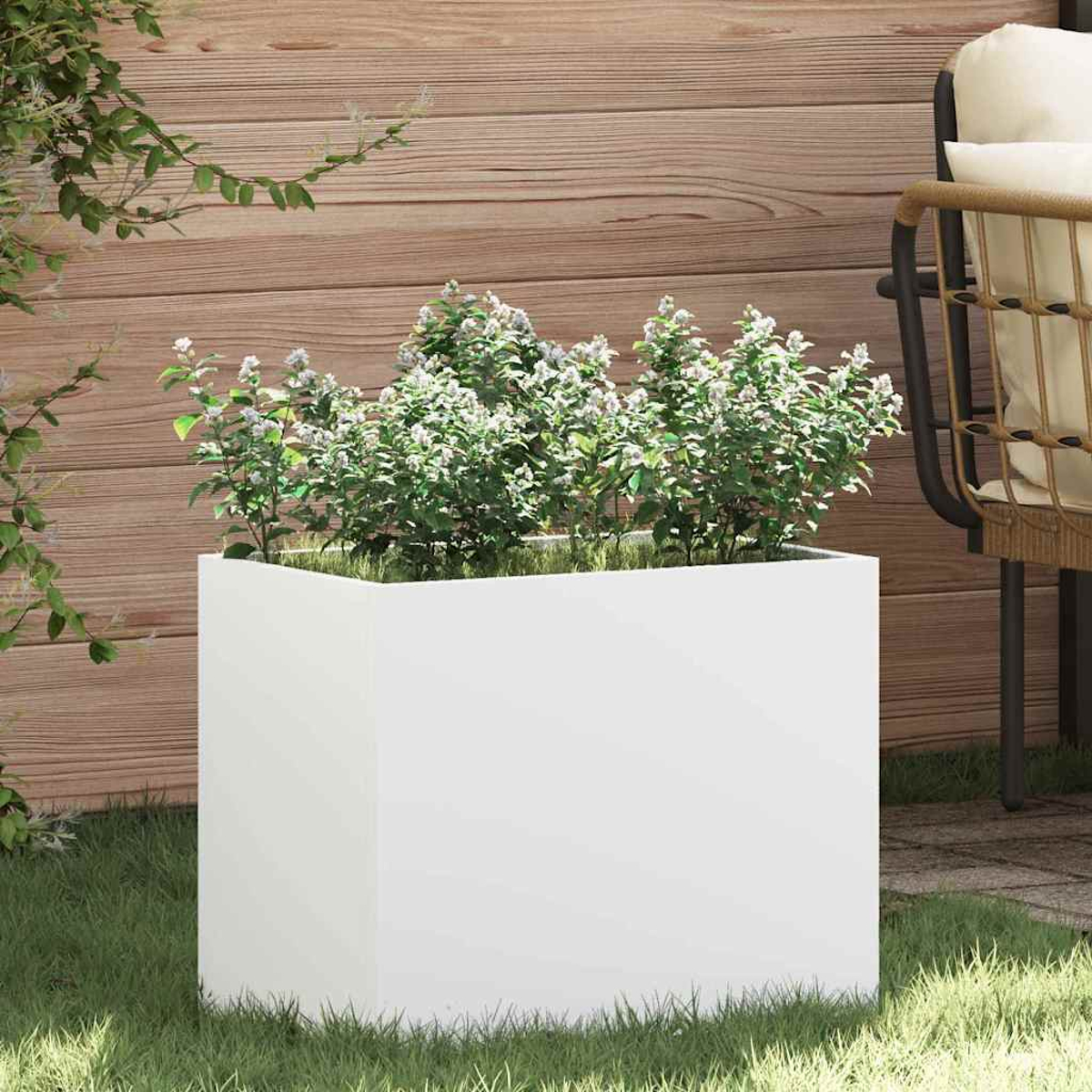 Ein weißer quadratischer Pflanzkübel mit grünen Pflanzen und weißen Blüten steht auf Gras. Eine Holzwand und ein Stuhl sind im Hintergrund.