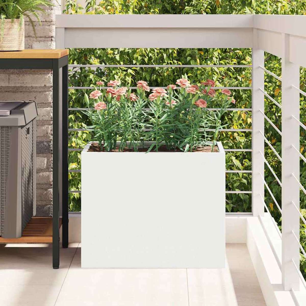 Weißer Pflanzkasten mit rosa Blumen auf einem Balkon. Grünes Laub im Hintergrund. Weißes Geländer rechts. Ziegelmauer links.