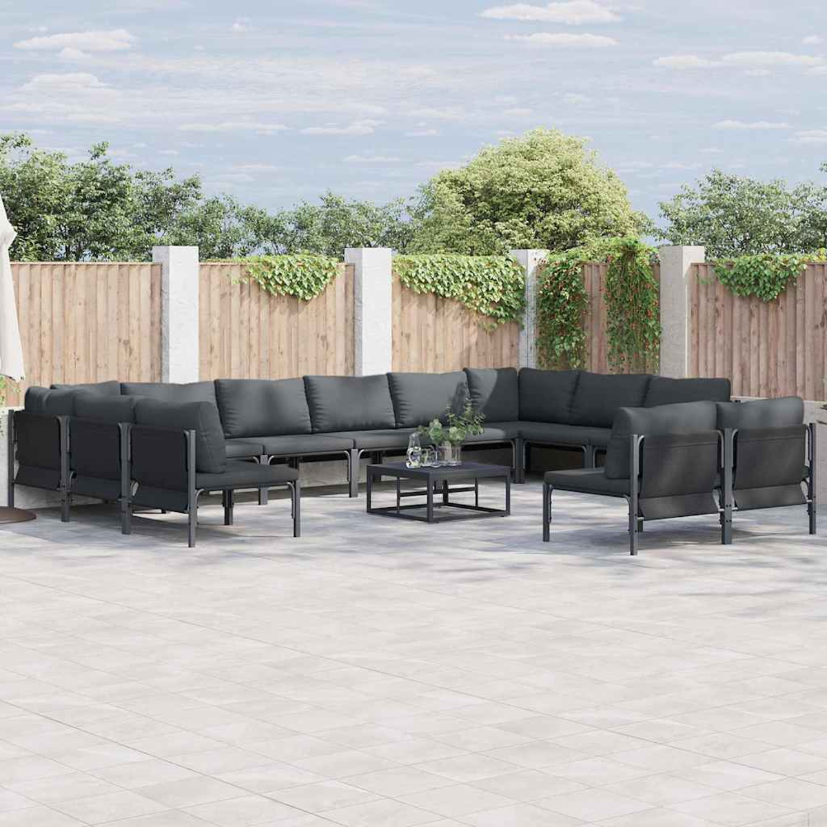 Outdoor-Sitzgruppe. Dunkelgraue Sofas und Stühle auf einer gefliesten Terrasse, kleiner schwarzer Tisch, Holzzaun, grünes Laub, bewölkter Himmel.