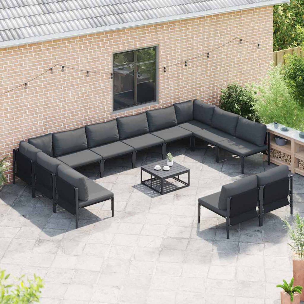 Terrassenmöbel-Set mit grauen Kissen, U-förmig angeordnet, mit kleinem Tisch. Backsteinmauer und Grün im Hintergrund.