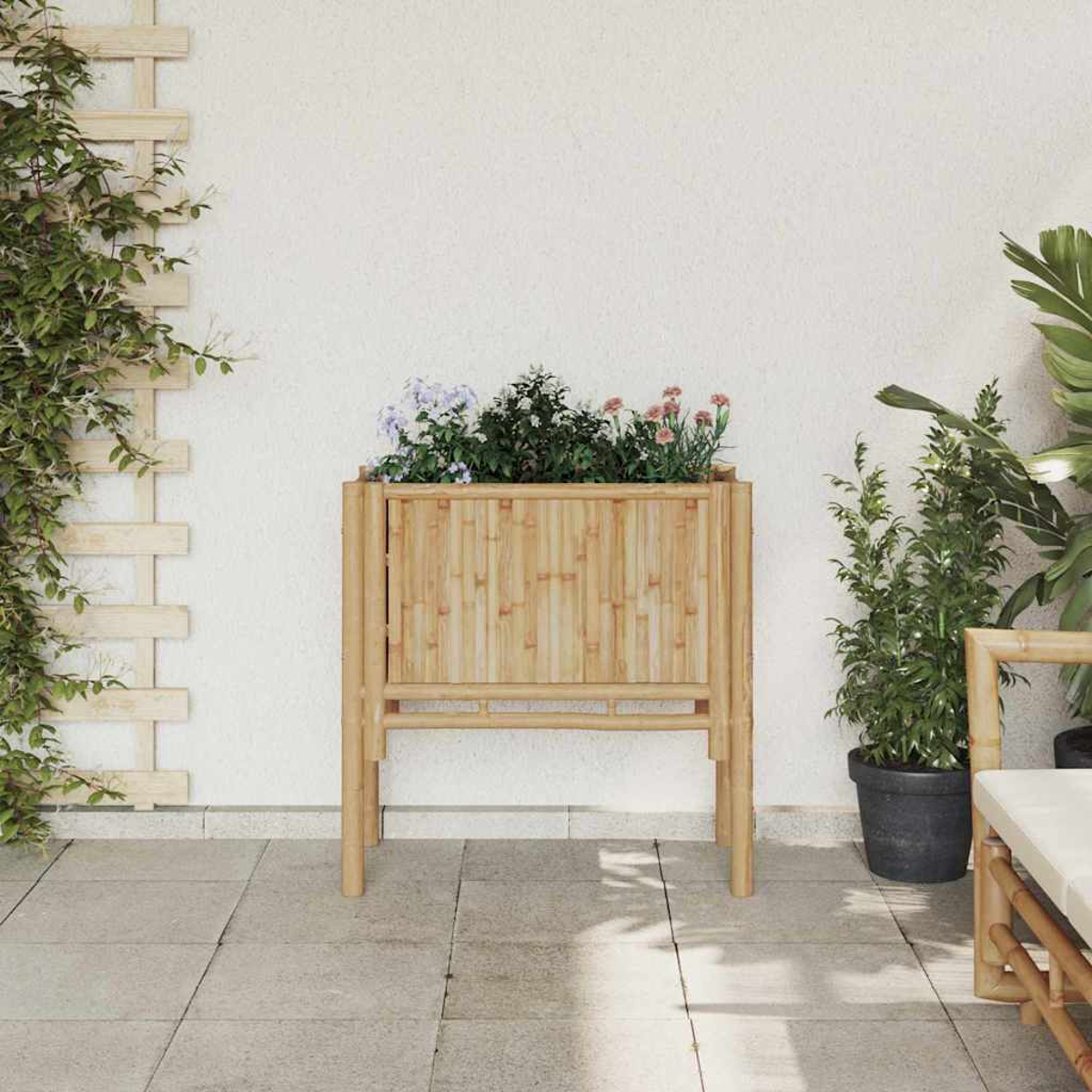 Una fioriera in bambù con fiori si trova su un patio. Le piante incorniciano la scatola e un muro bianco. Un traliccio con una pianta rampicante è a sinistra.