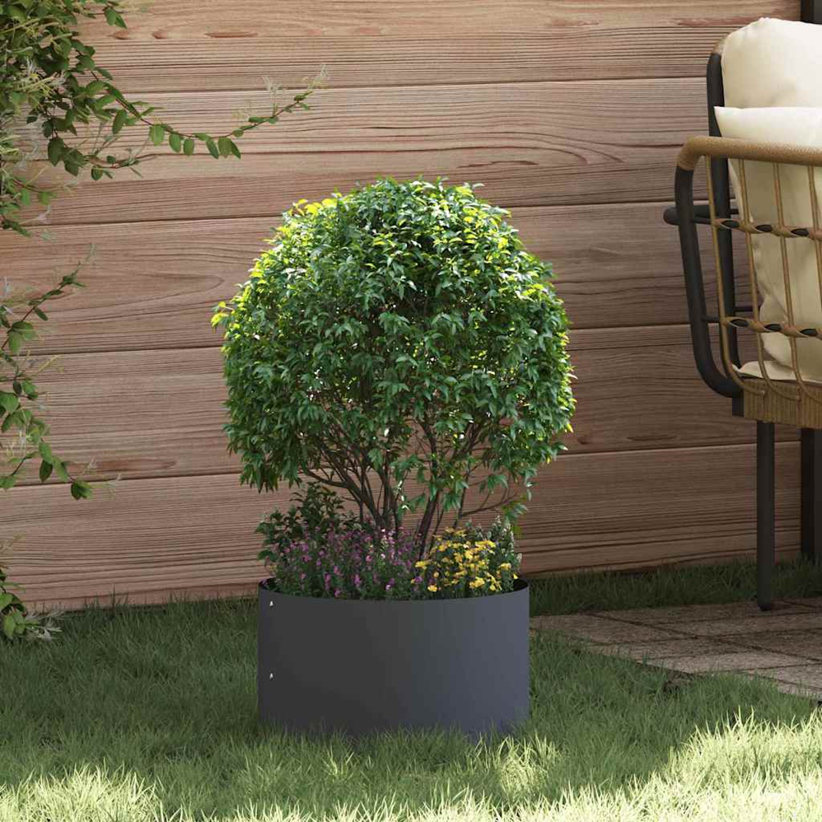 Un albero verde e frondoso in una fioriera grigio scuro. Dietro un muro di legno e una sedia. La scena è sull'erba.