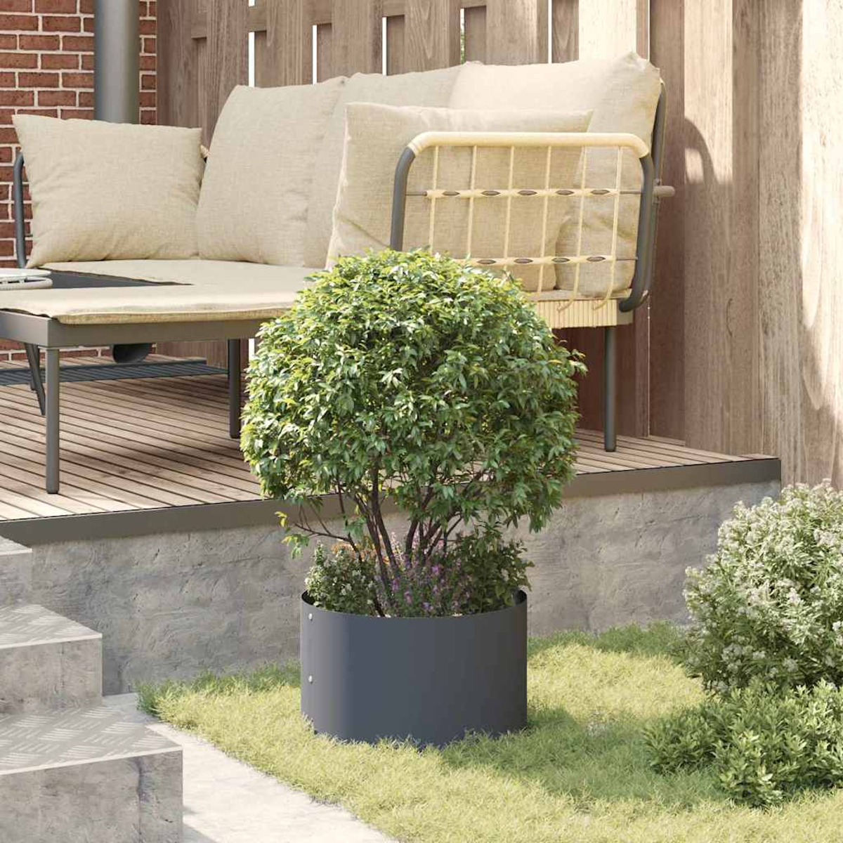 Un piccolo albero in una fioriera grigio scuro è sull'erba. Un set da patio con cuscini beige è dietro l'albero. Sono visibili delle scale.