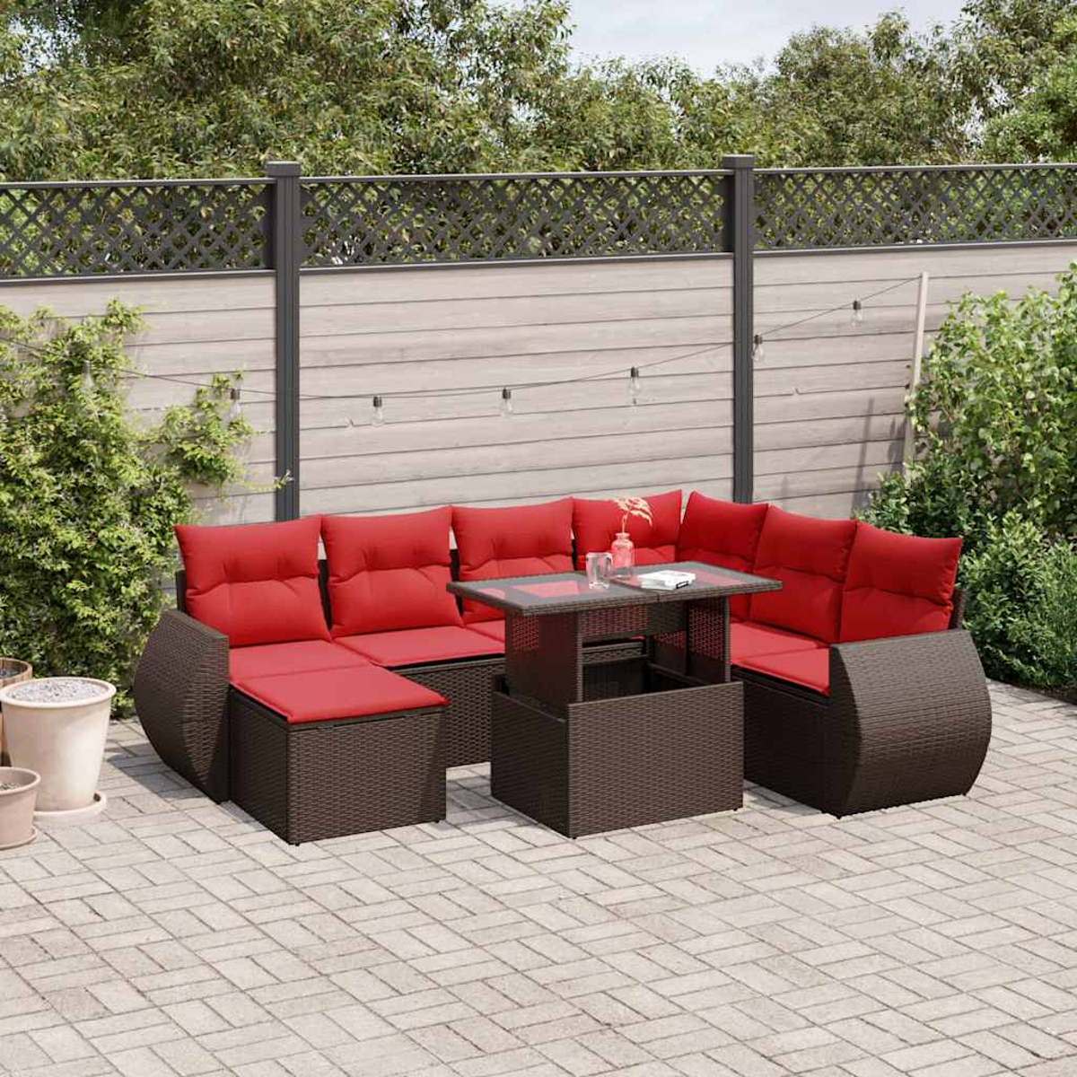 Outdoor-Korbsessel mit roten Kissen, Tisch mit Glasplatte. Auf einer Ziegelterrasse vor einem Holzzaun. Im Hintergrund grüne Pflanzen.
