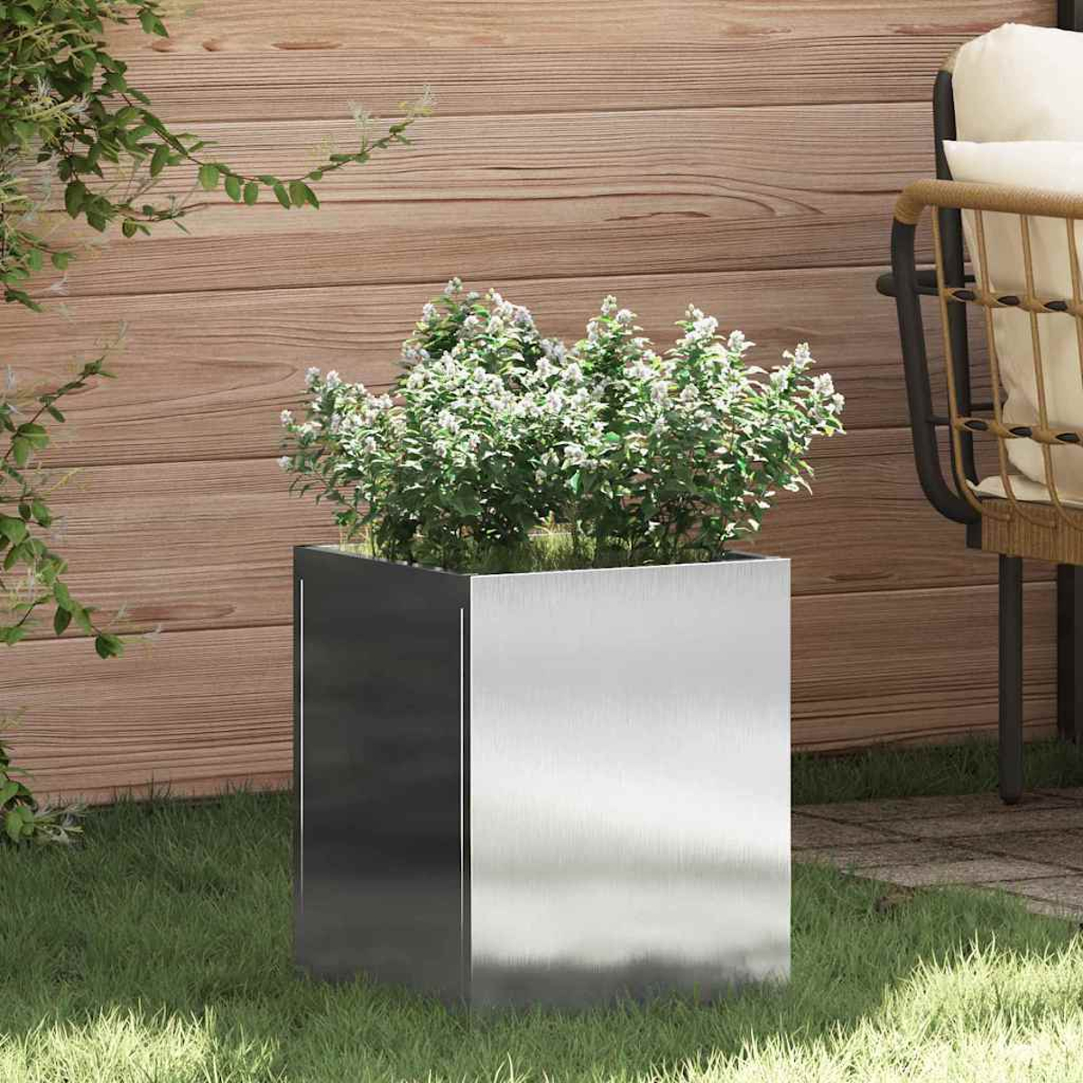 Ein quadratischer Metallpflanzer mit weißen Blumen und grünen Blättern auf Gras. Ein Holzzaun und ein Stuhl sind im Hintergrund.