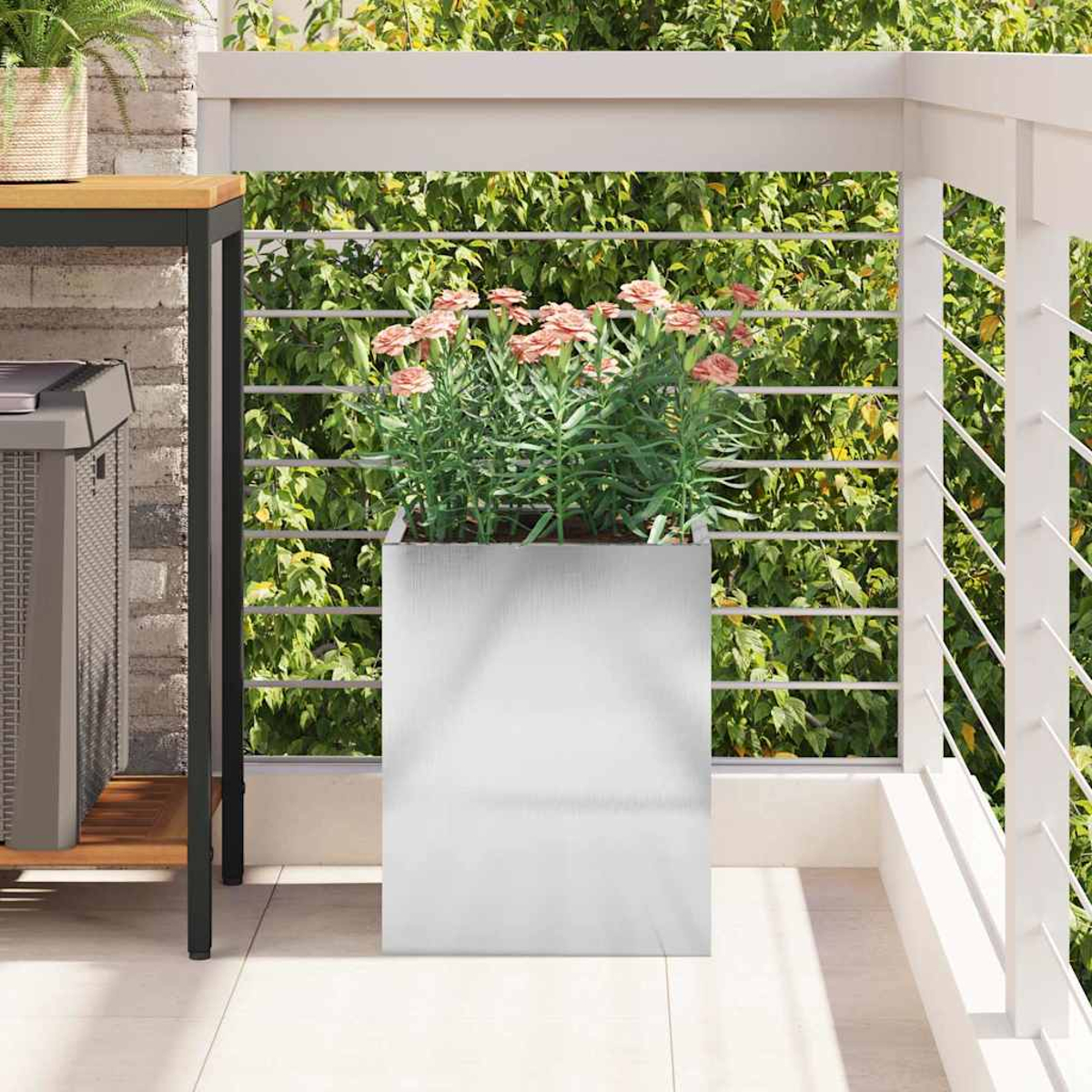 Ein silberner Pflanzkübel mit rosa Blumen auf einem Balkon. Ein weißes Geländer und grünes Laub im Hintergrund.