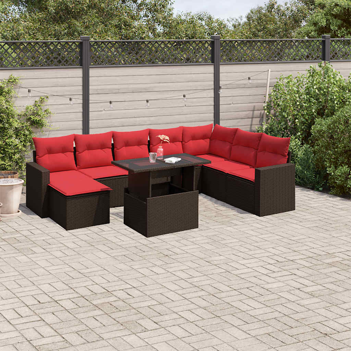 Eine Terrassenszene zeigt braune Korbmöbel mit roten Kissen, einen Tisch und einen gemusterten Ziegelboden. Grüne Büsche und ein Zaun befinden sich im Hintergrund.