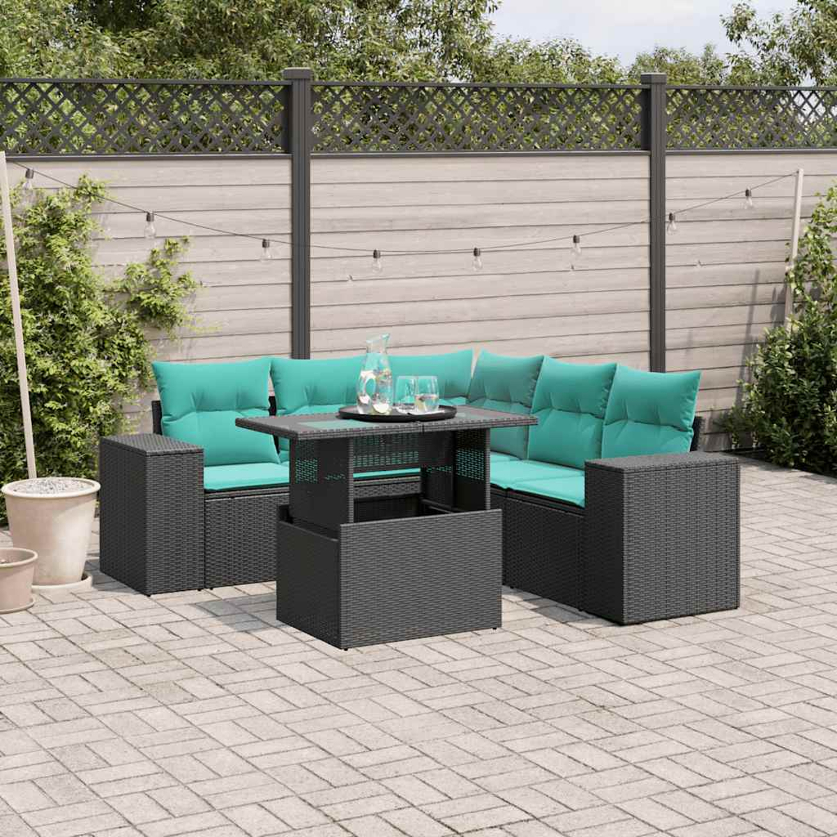 Terrassenset mit türkisfarbenen Kissen und grauen Korbmöbeln. Tisch mit Wasserkrug. Holzzaun im Hintergrund.