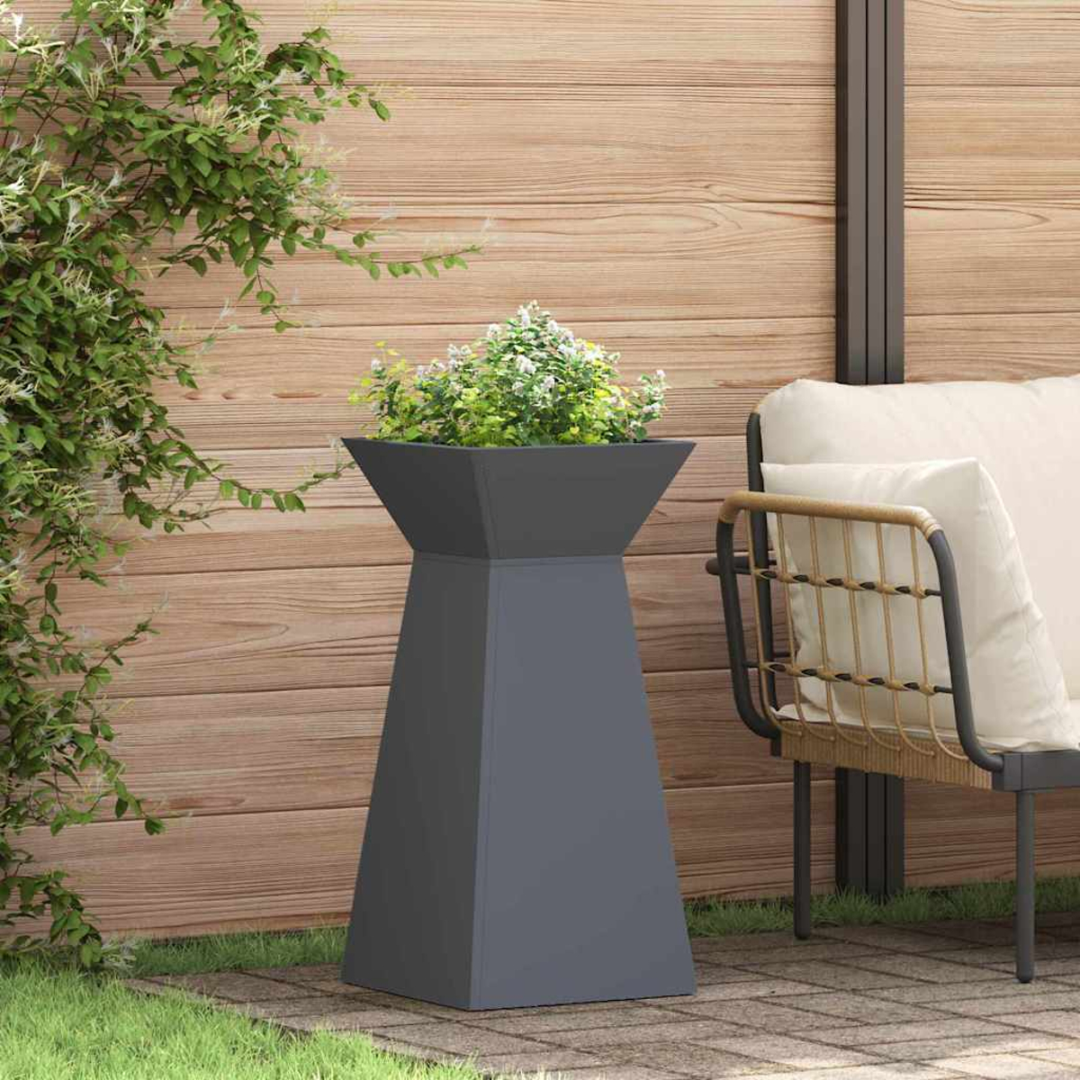 Una fioriera grigia con piante si trova su un patio, accanto a una sedia. Sullo sfondo una staccionata di legno e verde.