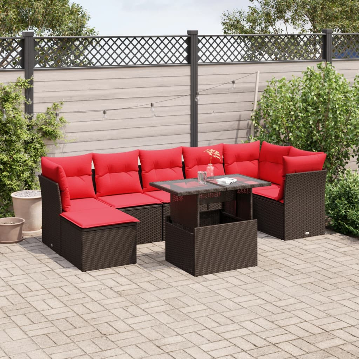 Gartenmöbel-Set mit roten Kissen und einem braunen Tisch. Auf einer Terrasse mit Zaun und Pflanzen im Hintergrund.
