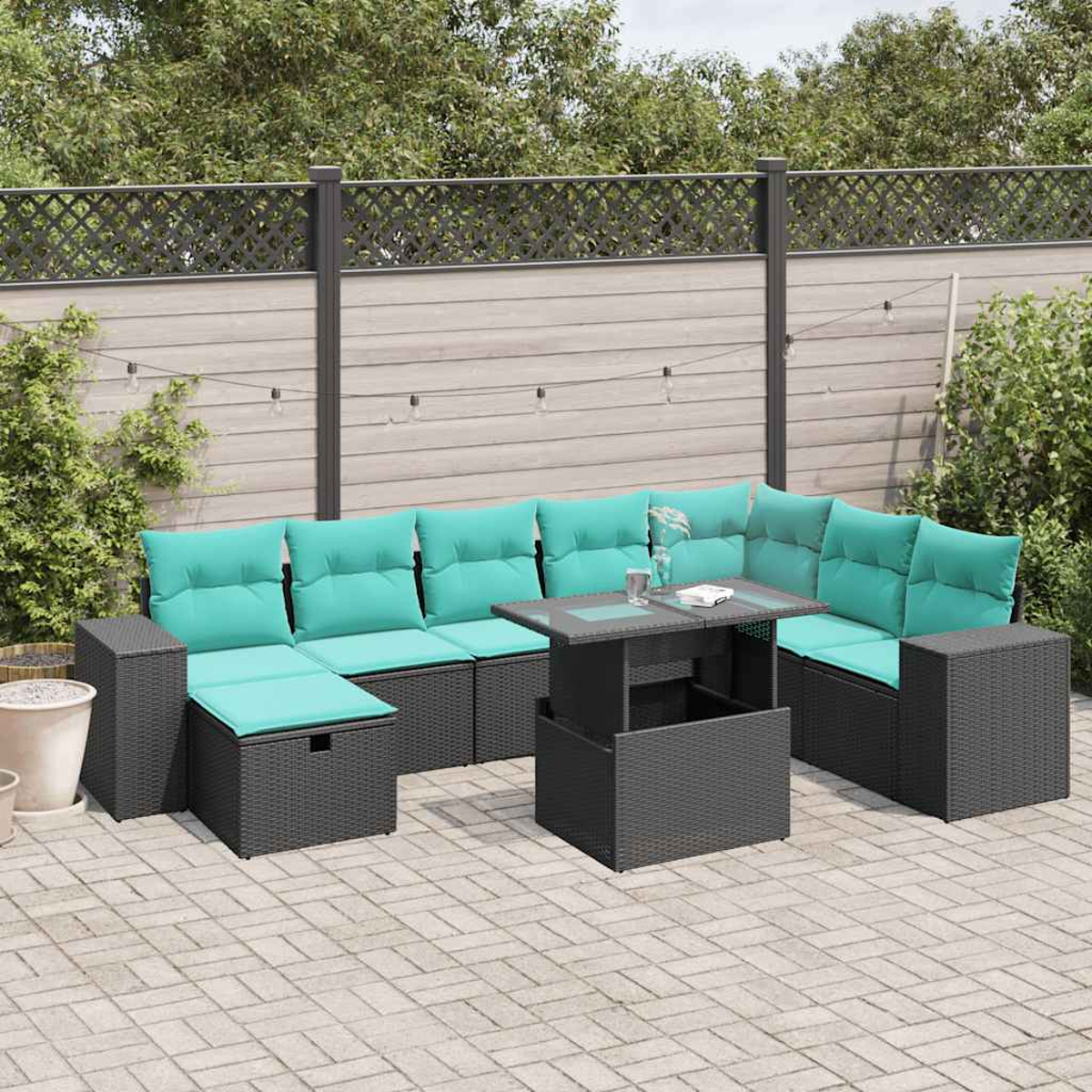 Outdoor-Sitzgruppe mit türkisfarbenen Kissen, einem Tisch und einem grauen Korbmuster. Auf einer Terrasse mit Zaun und Grün im Hintergrund.