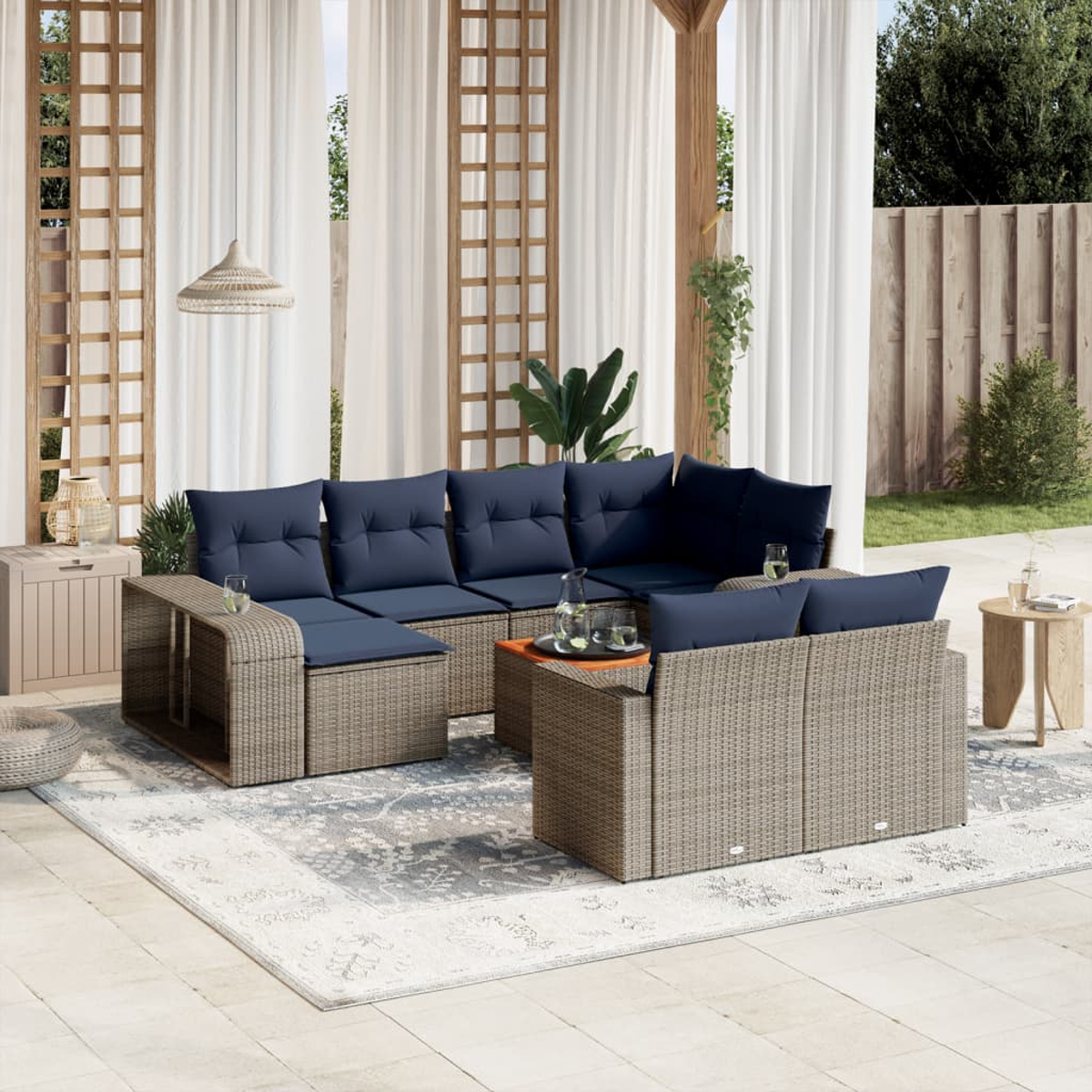 Outdoor-Sitzgruppe mit marineblauen Kissen auf einem gemusterten Teppich. Ein kleiner Tisch hält Getränke. Weiße Vorhänge und Holzgitter im Hintergrund.