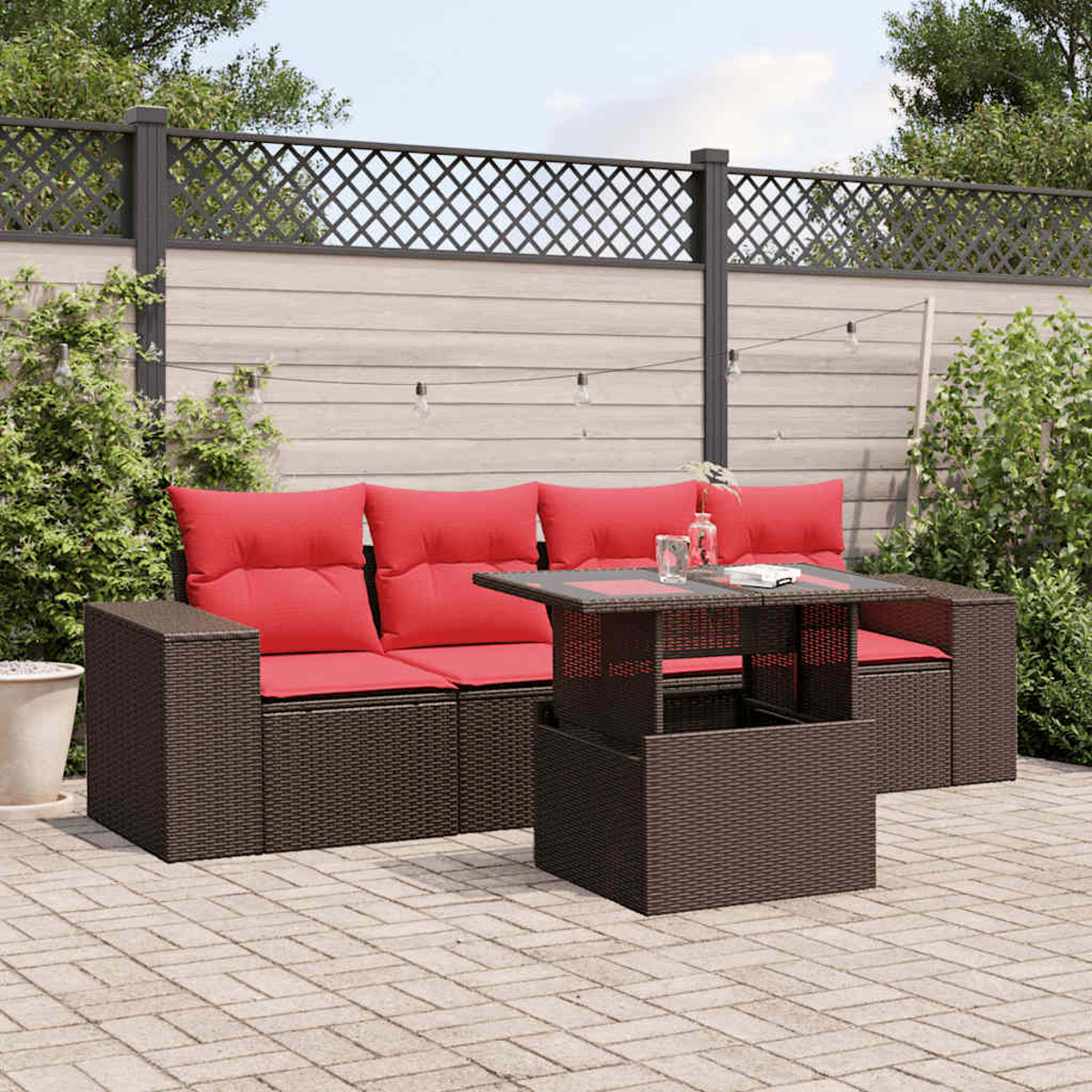 Garten-Set mit roten Kissen und Glastisch. Im Hintergrund ein Zaun und Pflanzen. Lichterketten hängen darüber.