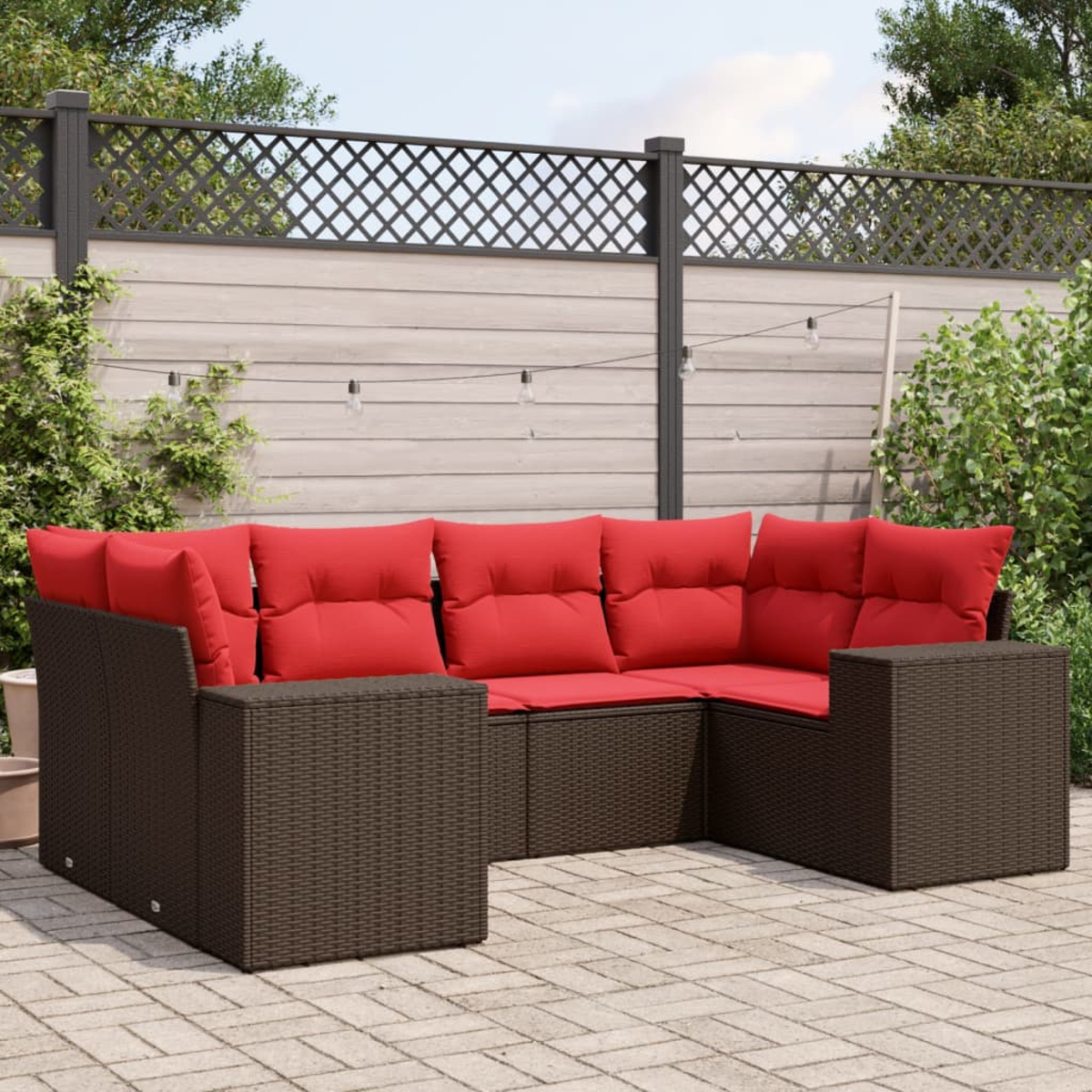 Outdoor-Ecksofa mit roten Kissen auf einer Steinpflasterung. Dunkelbrauner gewebter Rahmen. Hintergrund: Zaun, Grün und Lichterketten.