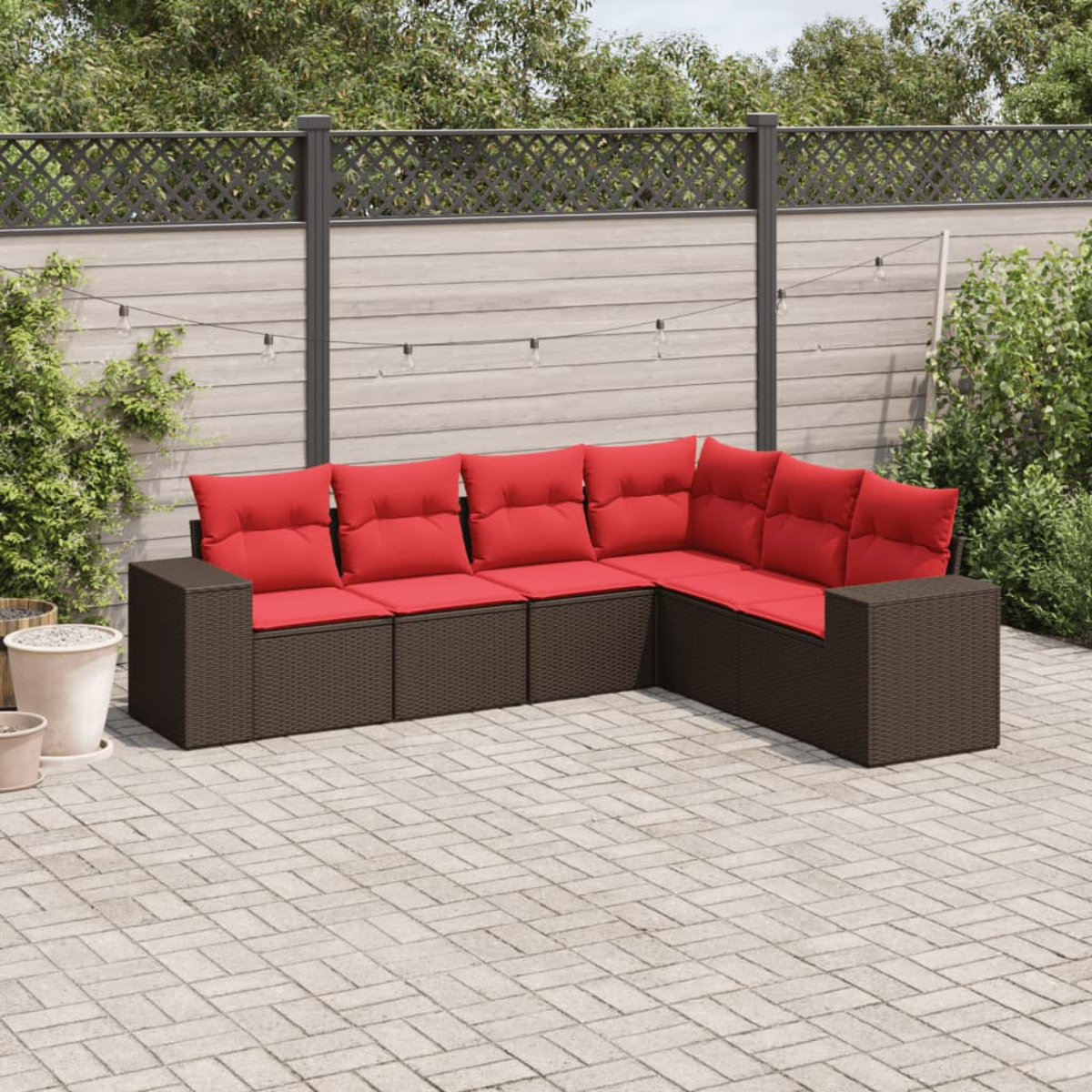 Outdoor-Ecksofa mit roten Kissen auf einer gepflasterten Terrasse. Brauner Korbrahmen. Zaun und Pflanzen im Hintergrund.
