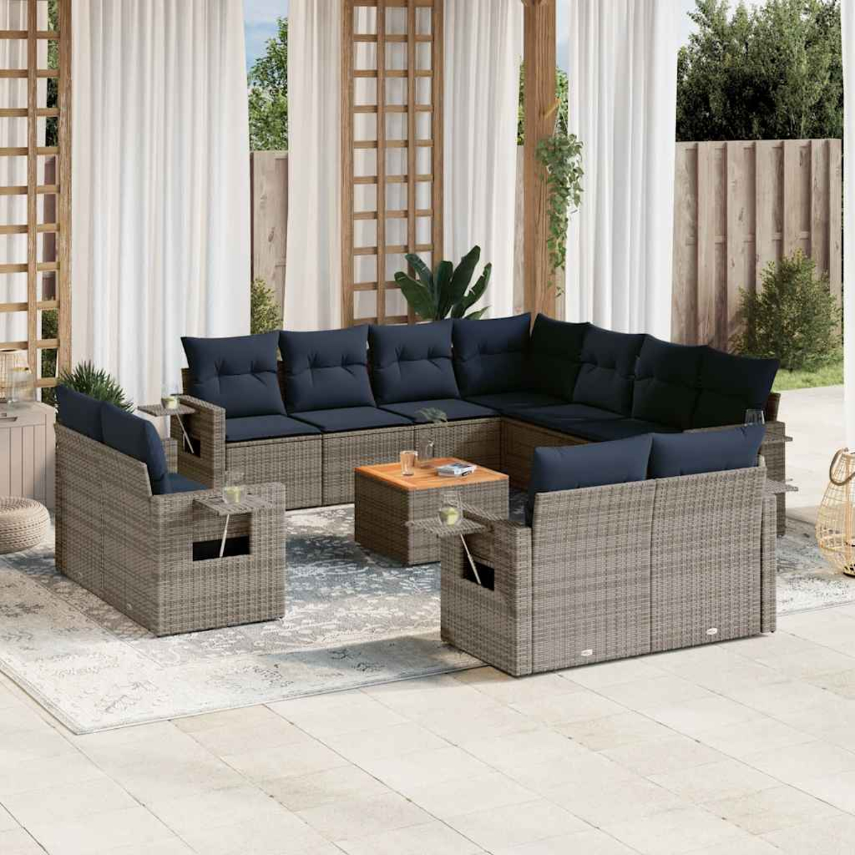 Terrassenmöbel-Set mit grauem Korbmöbel, marineblauen Kissen und einem Couchtisch aus Holz. Weiße Vorhänge und Grünzeug im Hintergrund.