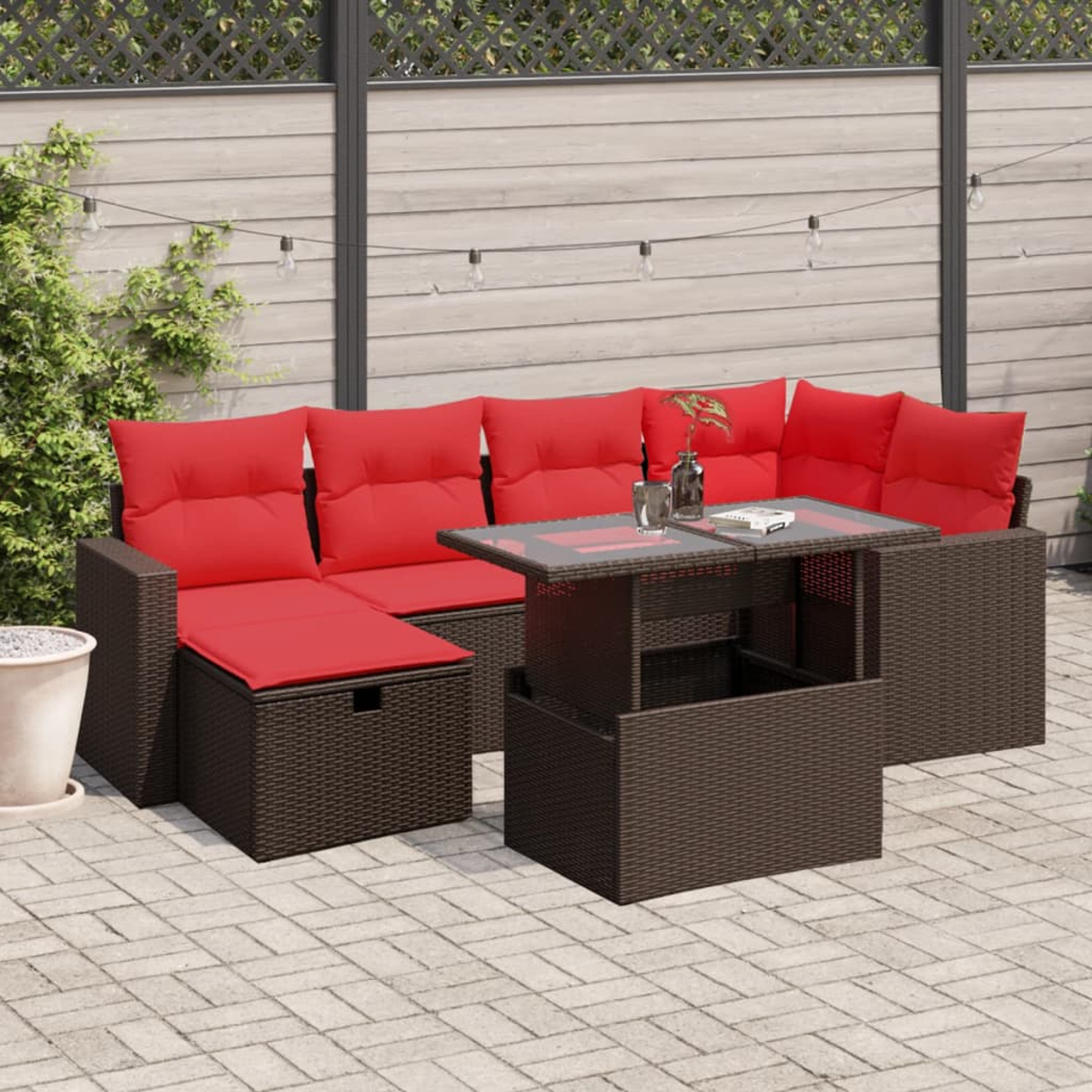 Outdoor-Ecksofa mit roten Kissen und einem Tisch. Der Hintergrund zeigt einen Zaun, Lichterketten und eine Pflanze. Die Terrasse ist gepflastert.