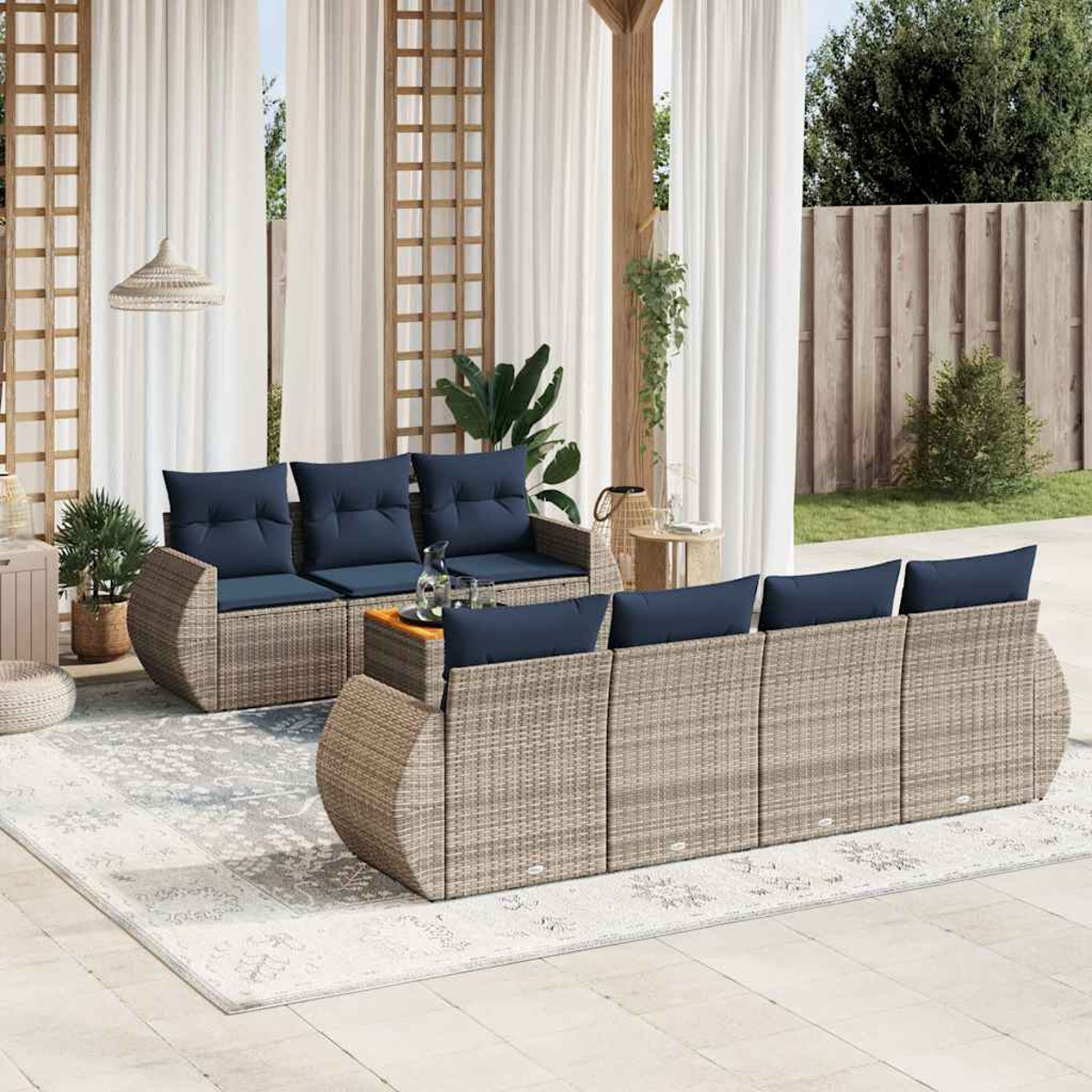 Outdoor-Sitzarrangement mit Korbsesseln, blauen Kissen und einem Holztisch auf einem gemusterten Teppich. Weiße Vorhänge und ein Holzzaun sind im Hintergrund.