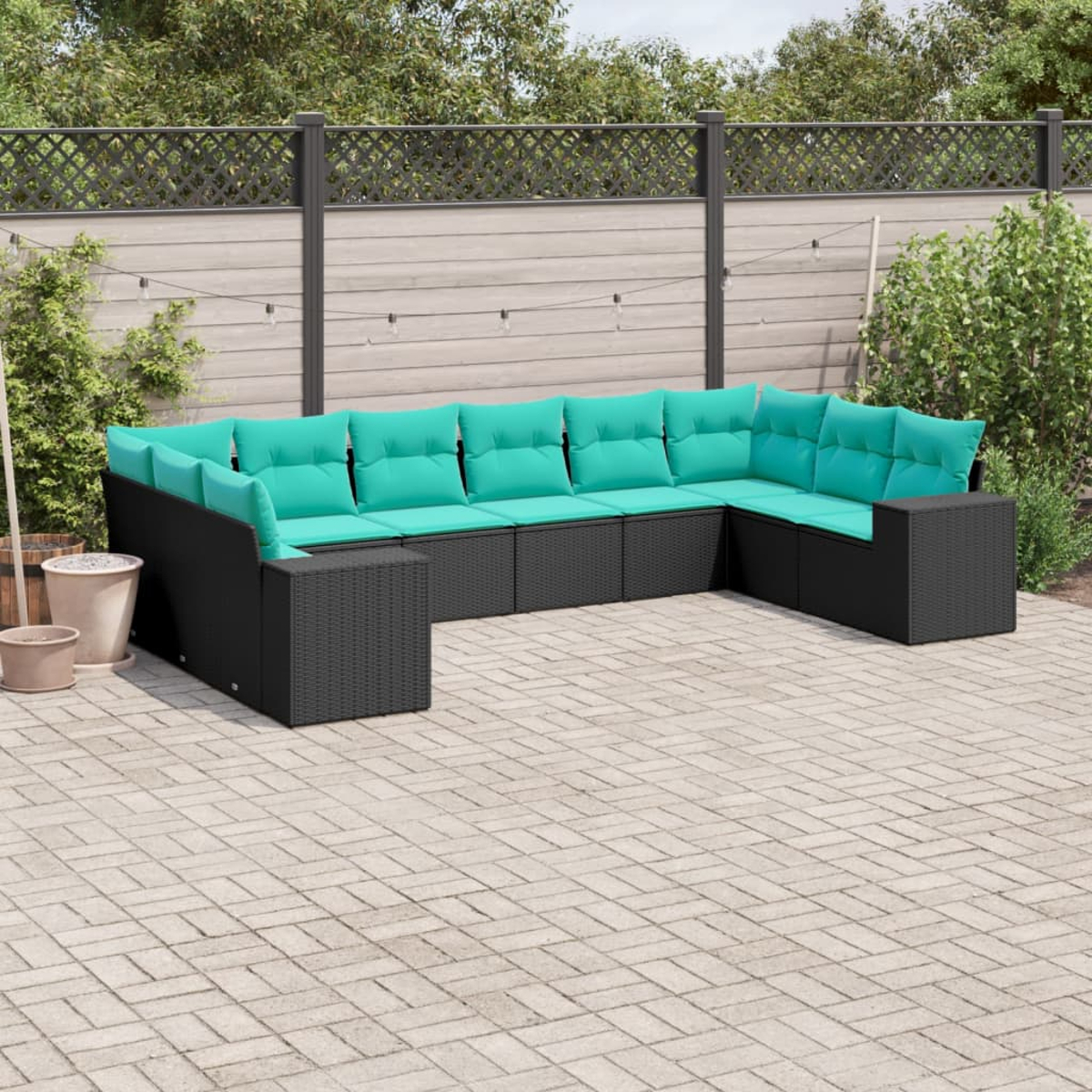 Ein schwarzes Outdoor-Ecksofa aus Korbgeflecht mit türkisfarbenen Kissen steht auf einer Steinterrasse. Der Hintergrund umfasst einen Zaun und Grün.